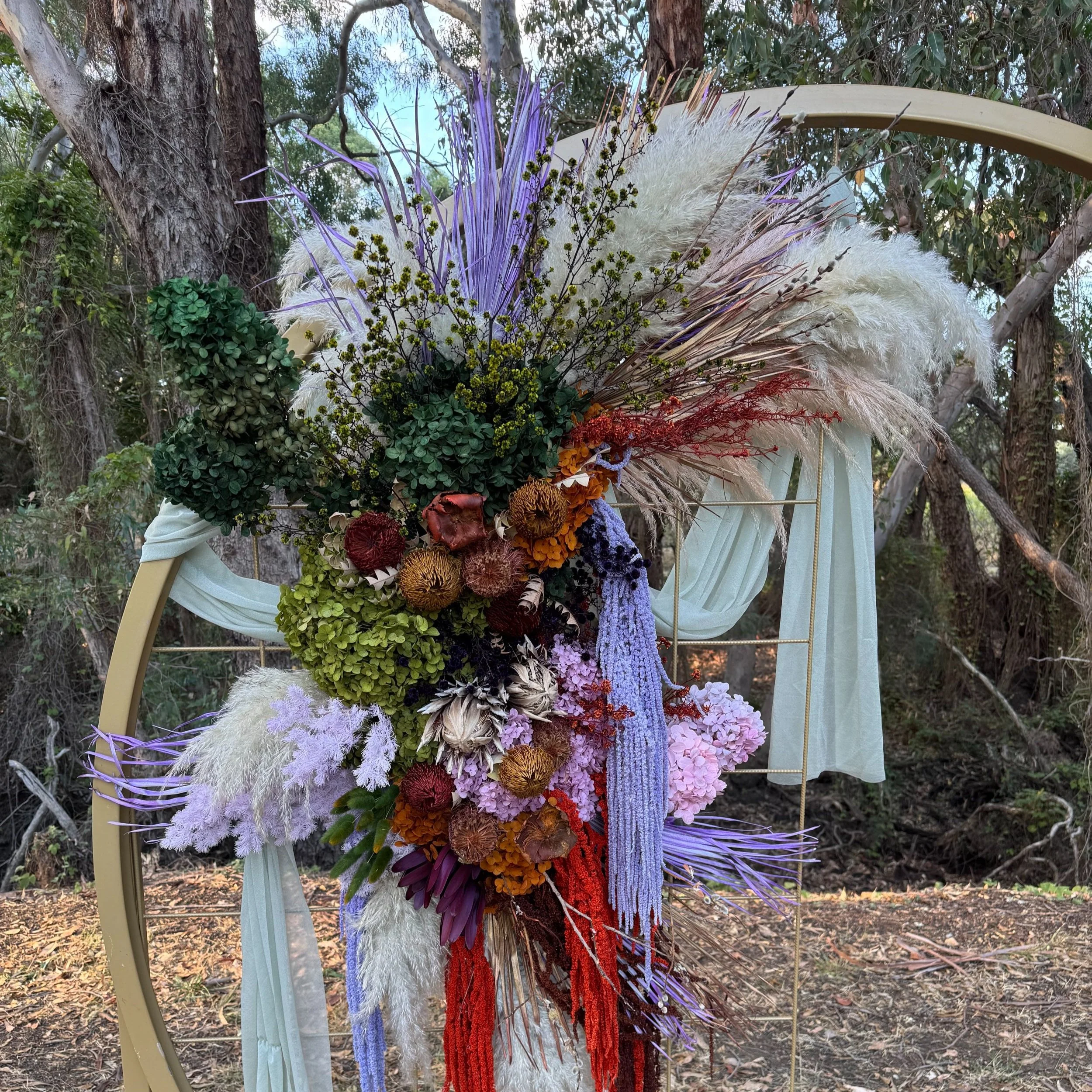 A floral installation with various colorful dried and preserved flowers and grasses, arranged on a curved metal frame with white fabric drapes, set outdoors with trees in the background.