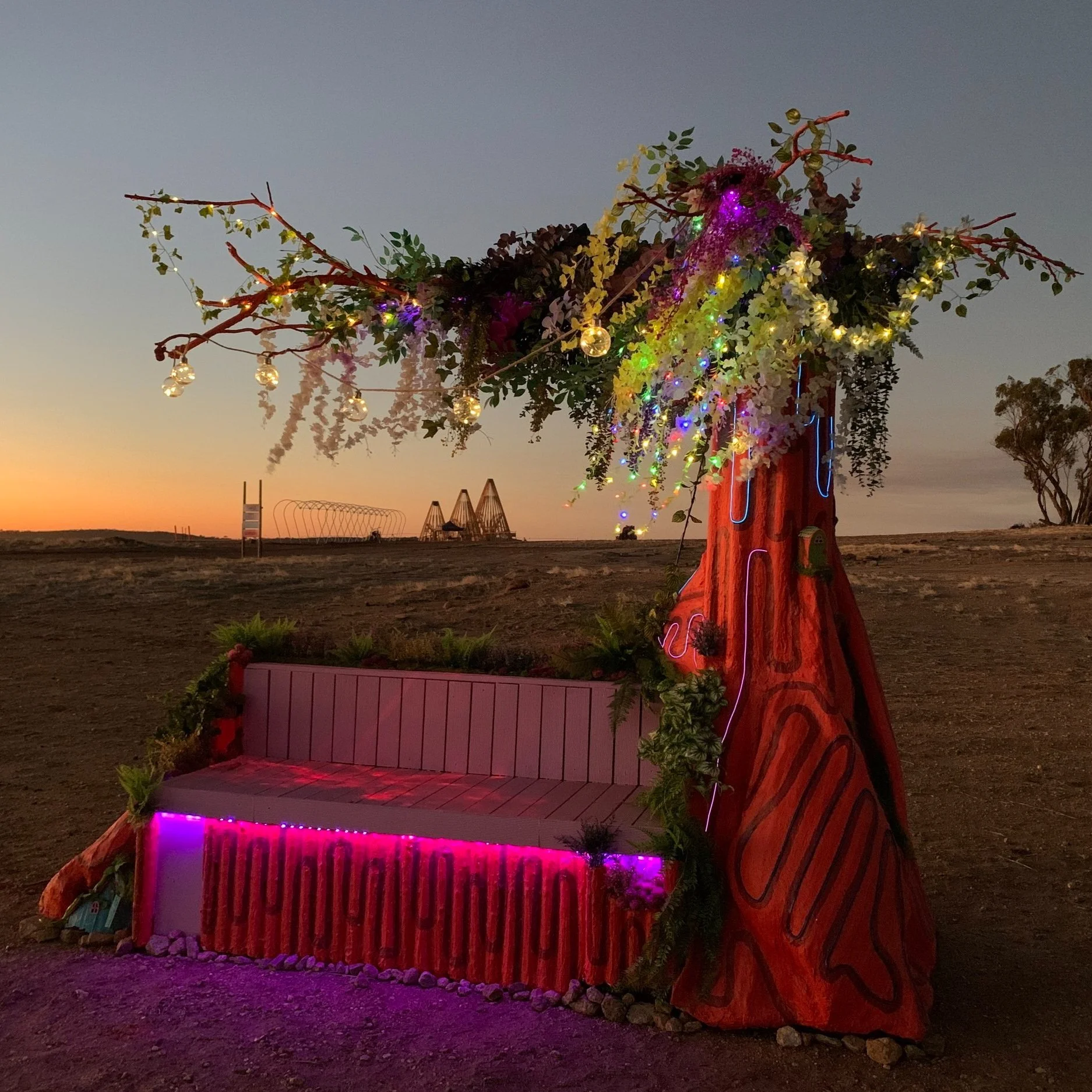 Decorated tree with colorful lights and flowers, and a pink bench underneath, in an open outdoor area during sunset.