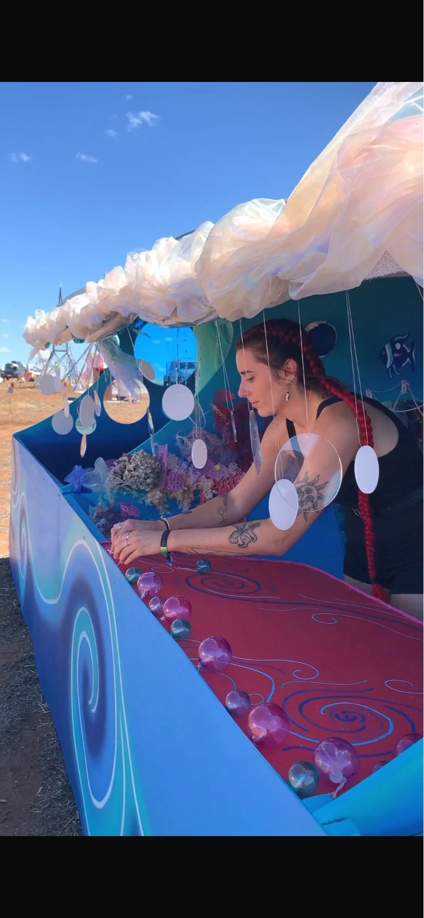 A woman with red braided hair and tattoos leaning into a colorful booth decorated with flowers, glass spheres, and a whimsical canopy, under a clear blue sky.