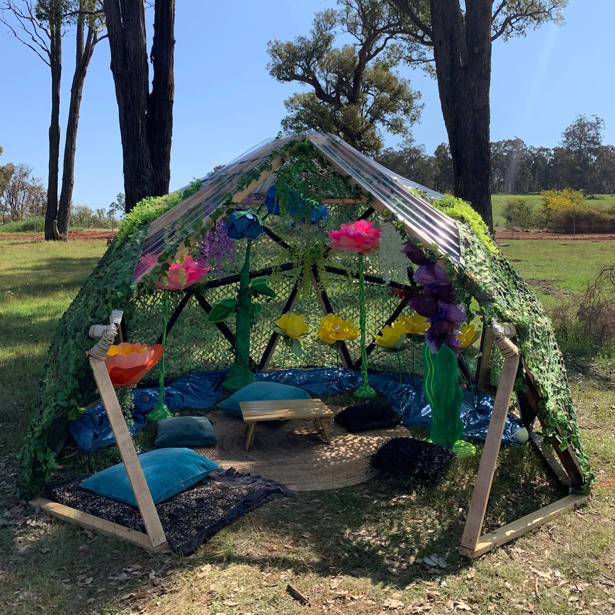 Colorful homemade fairy-tale-like outdoor shelter with pillows, a small table, and decorative flowers, set in a grassy park with trees and a bright blue sky in the background.
