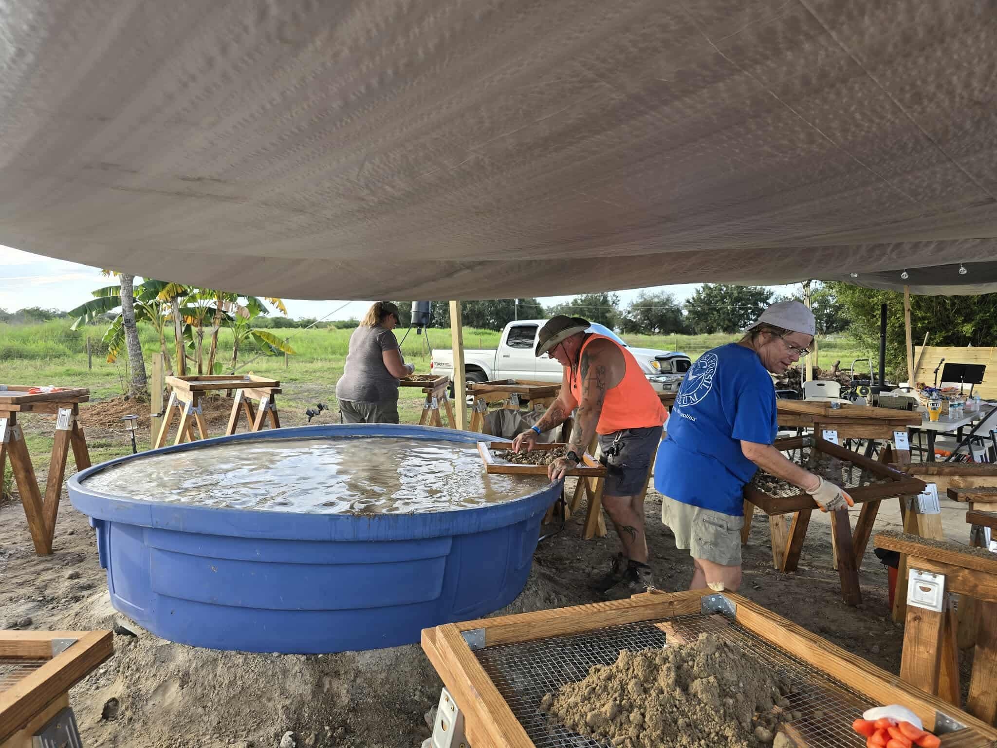 People working on a large outdoor project under a canopy, with a big blue water tank and wooden frames around. They are handling small objects or materials, with greenery and a pickup truck in the background.