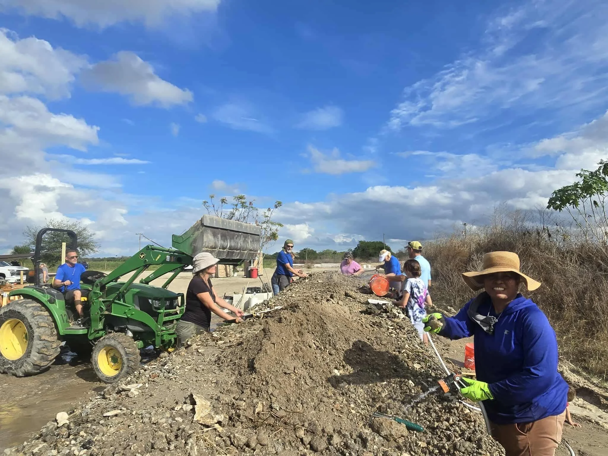 Group of people working outdoors on a construction or excavation site under a blue sky, some using tools and a tractor for digging or moving earth.