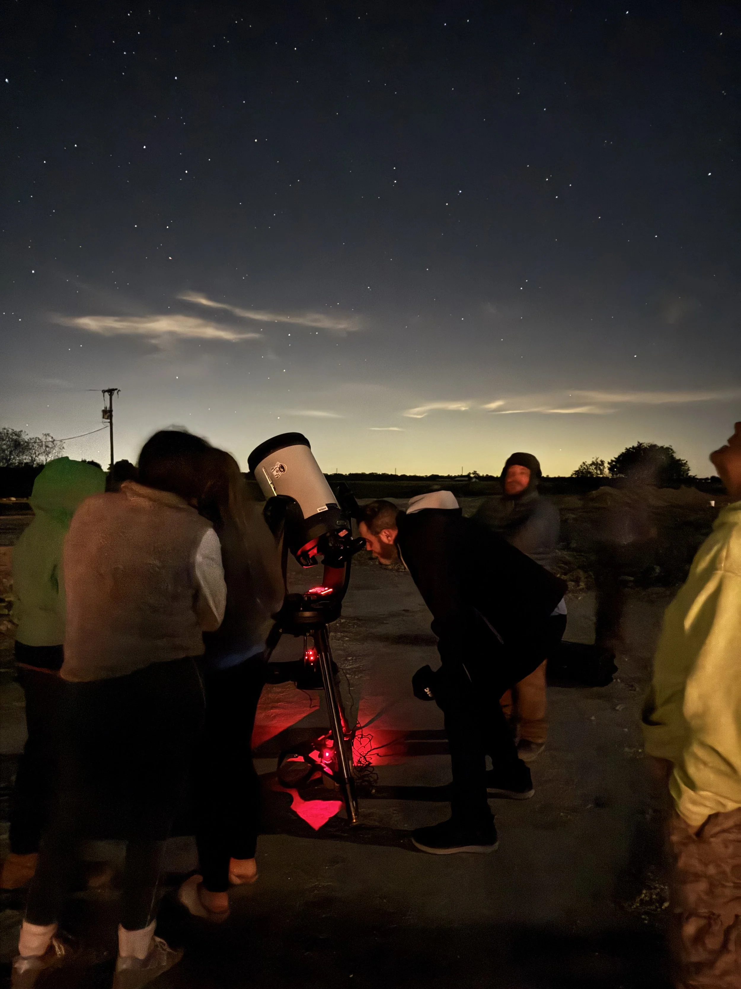 Group of people observing the night sky using a telescope outdoors at dusk or night.