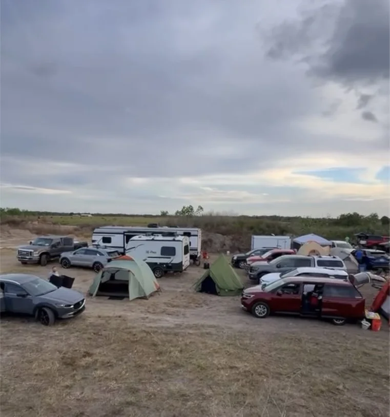 A campsite with multiple tents, RVs, and cars set up on a dirt field under a cloudy sky.