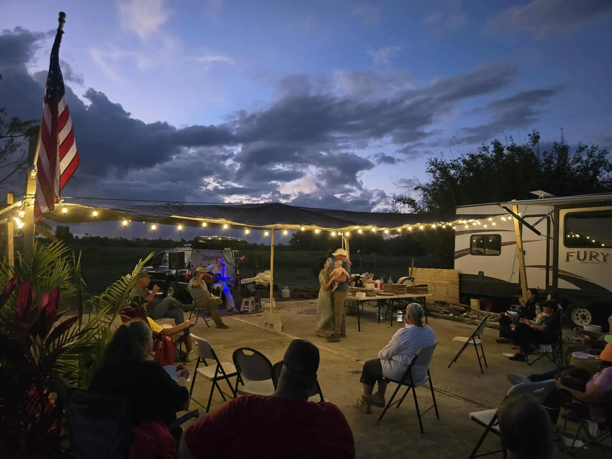 An outdoor gathering during dusk with string lights, an American flag, and people sitting on chairs, some facing a small stage with two people embracing, in front of a trailer and open field, under cloudy sky.