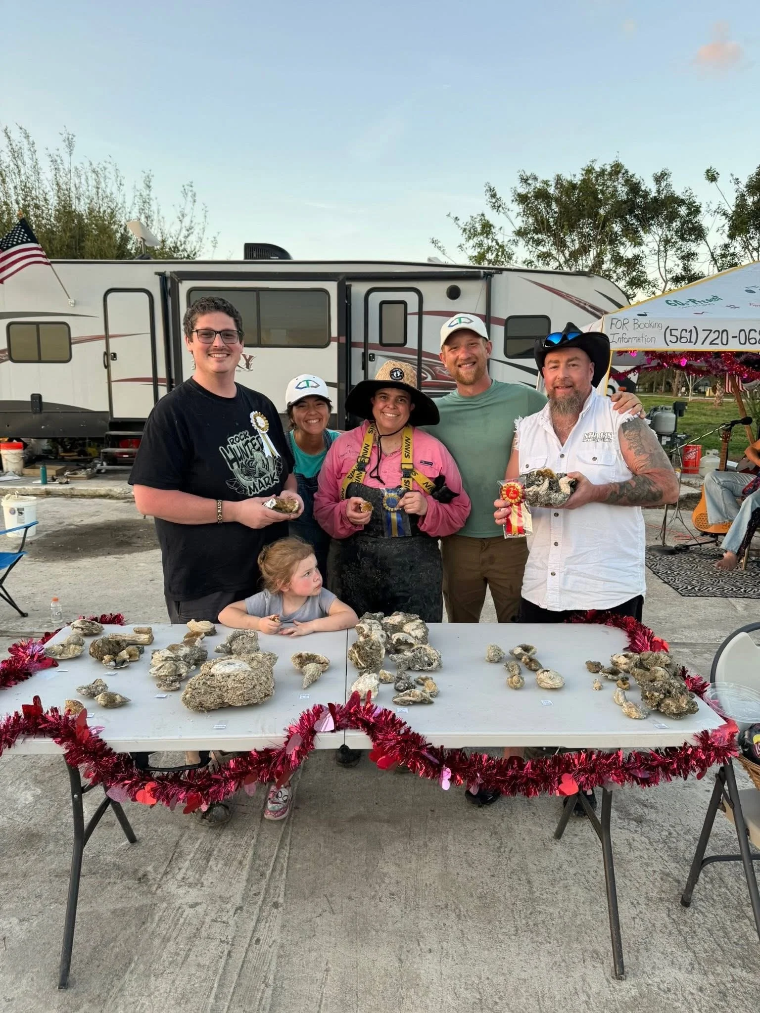 A group of six people and a young girl standing behind a table with various rocks or shells at an outdoor event, with RVs and trees in the background.