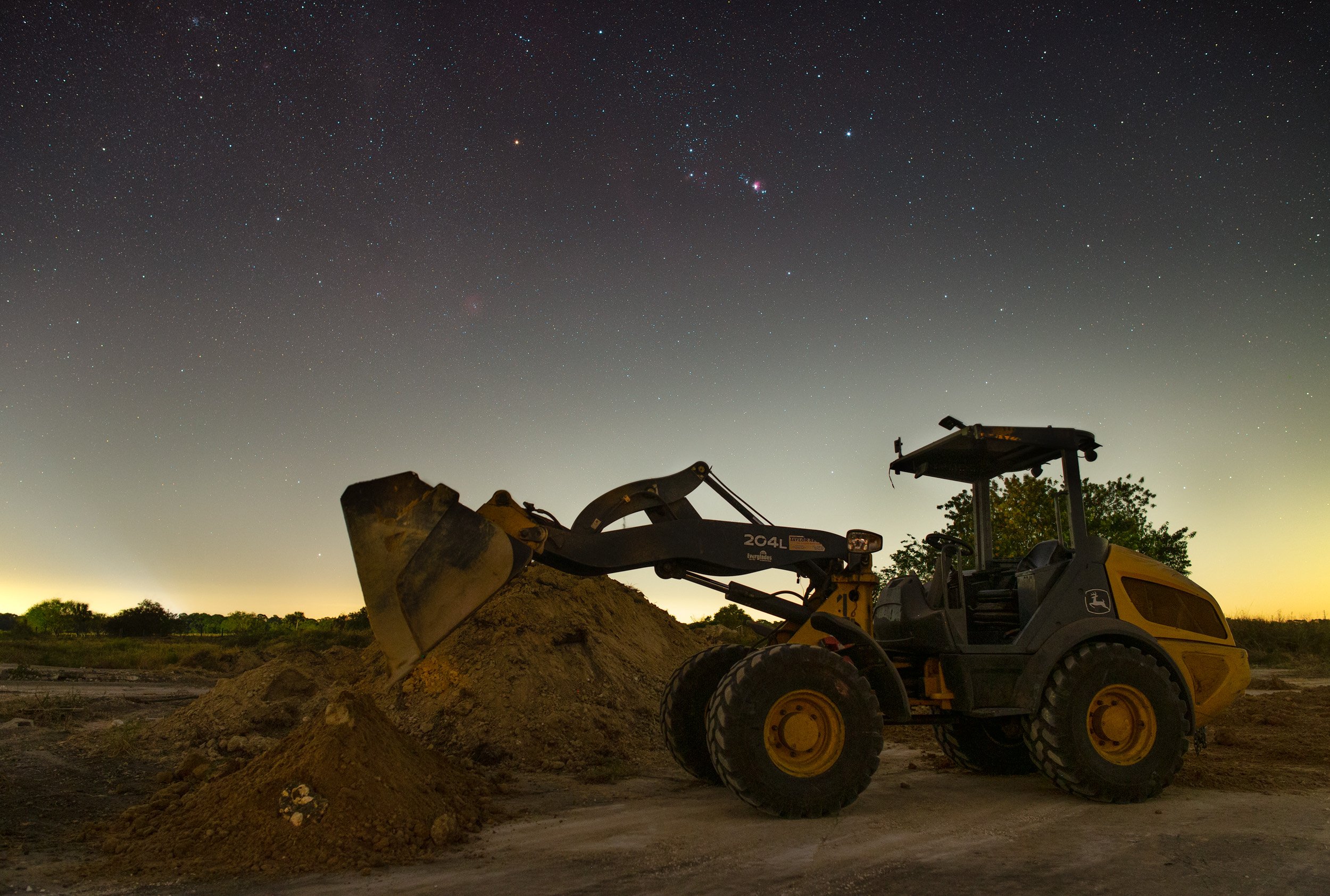 Construction vehicle, likely a backhoe loader, on a dirt construction site at night with a starry sky overhead.