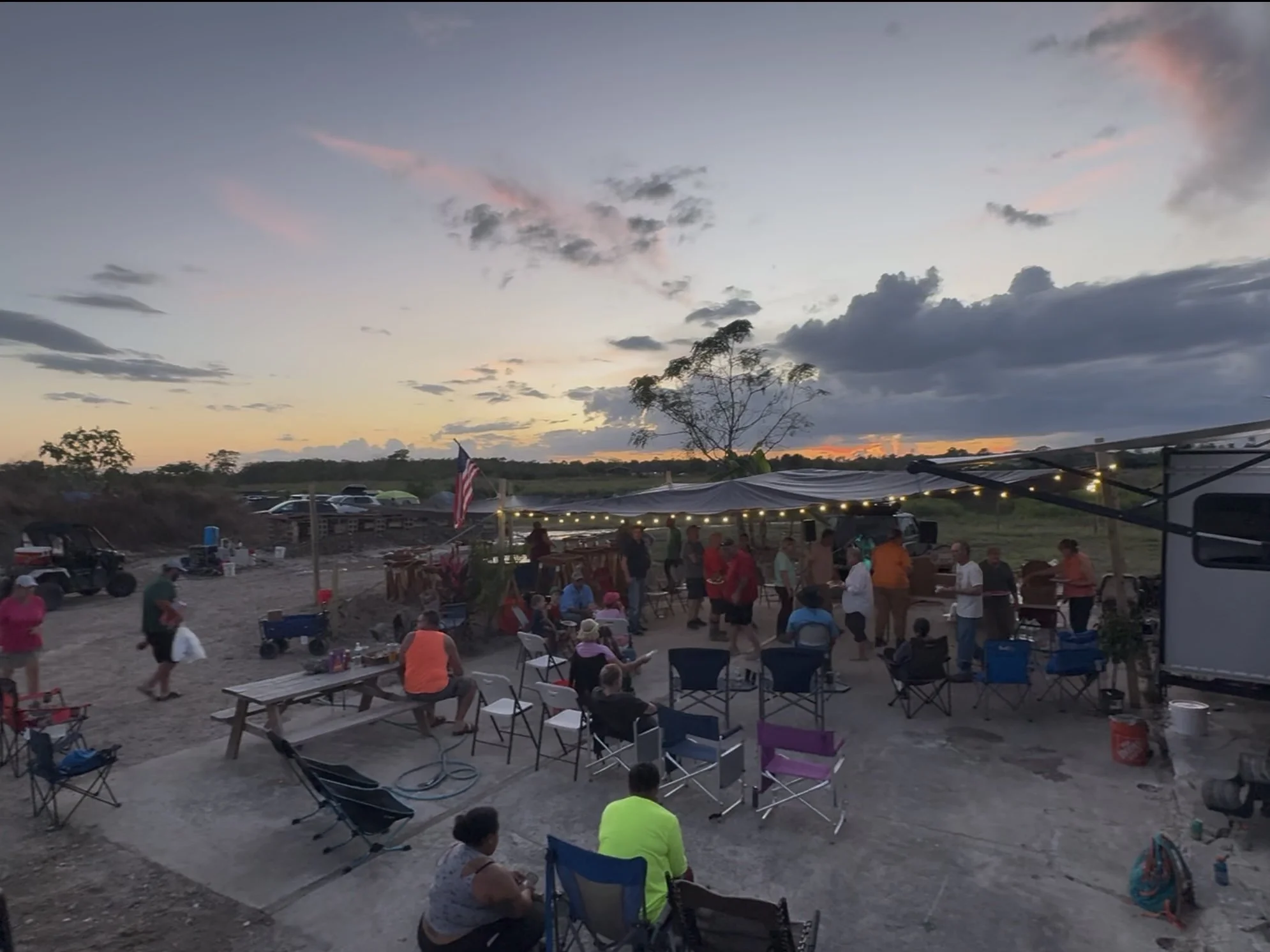An outdoor gathering at sunset with people socializing under a canopy decorated with string lights. There are chairs, tables, and a few vehicles, including a golf cart, in an open area with a scenic sunset sky.