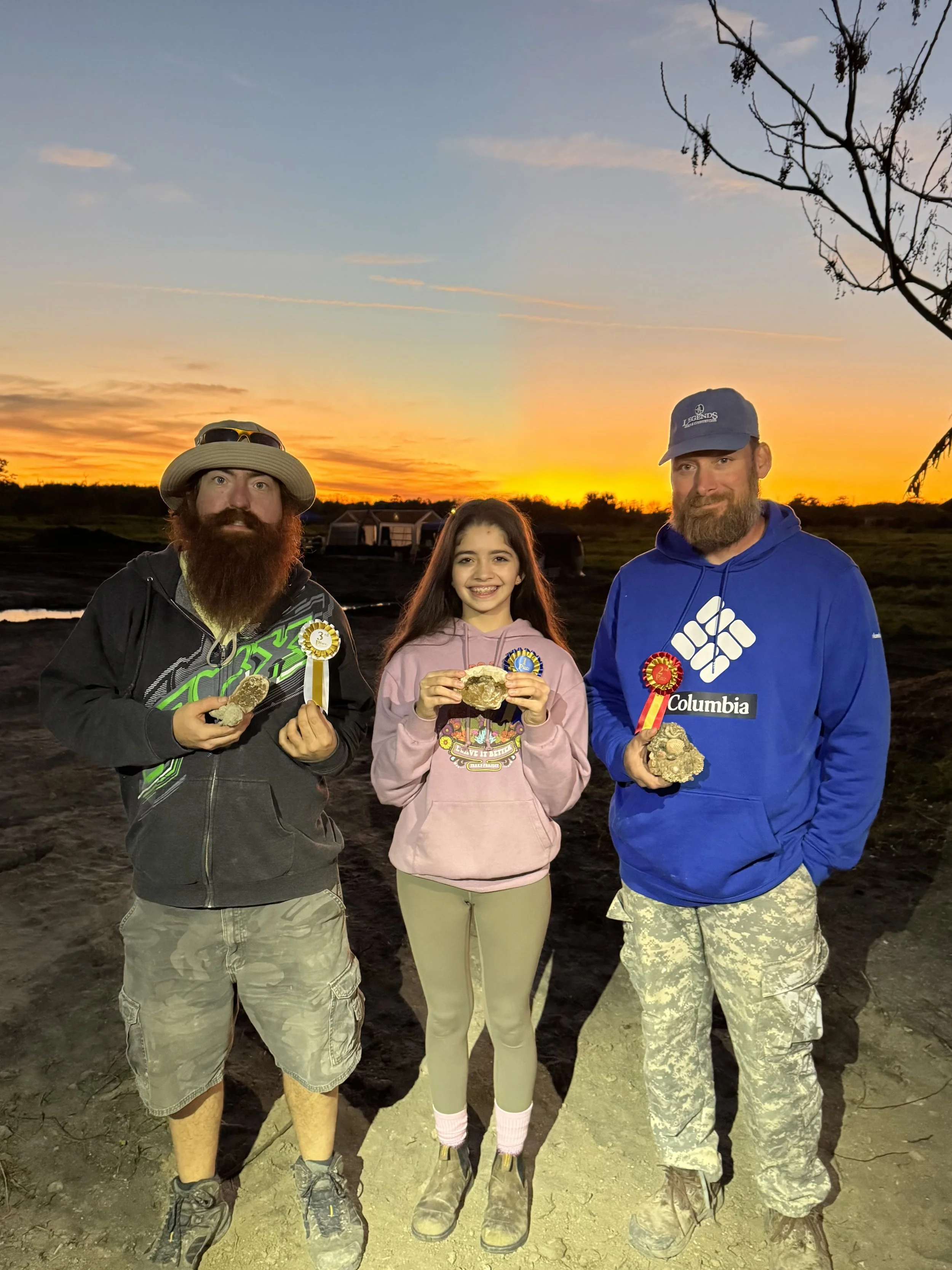 Three people, two men and a young girl, standing outdoors at sunset, holding rocks and wearing medals and casual clothing.