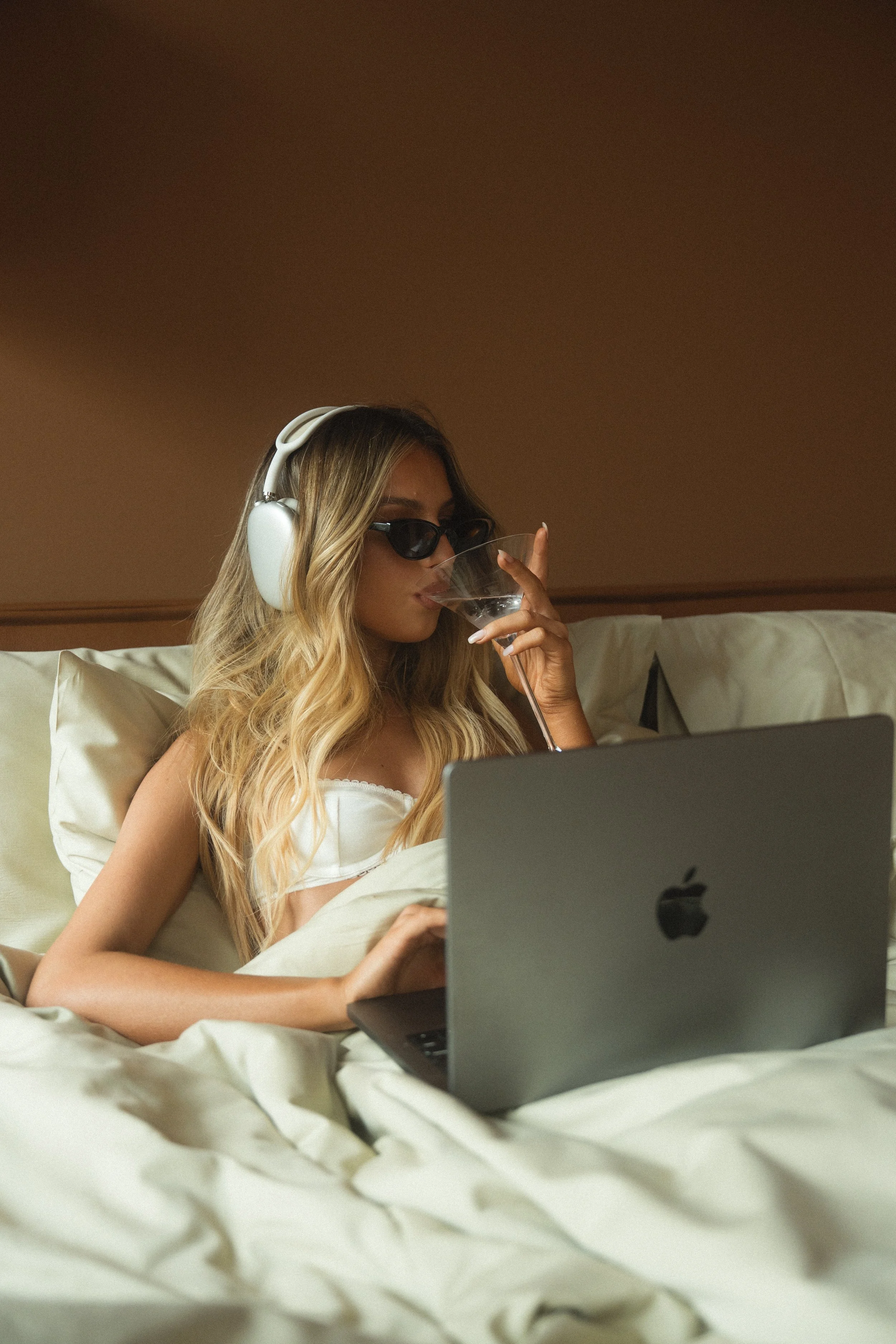 A woman lounging in bed, wearing sunglasses and headphones, drinking from a wine glass, using a silver MacBook