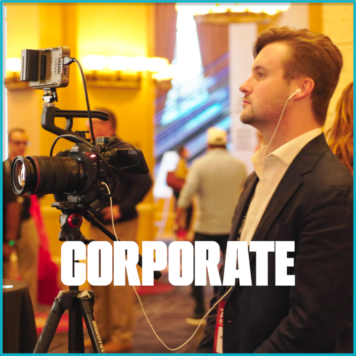 A man in a suit operating a professional video camera at a corporate event or conference, with several people visible in the background.