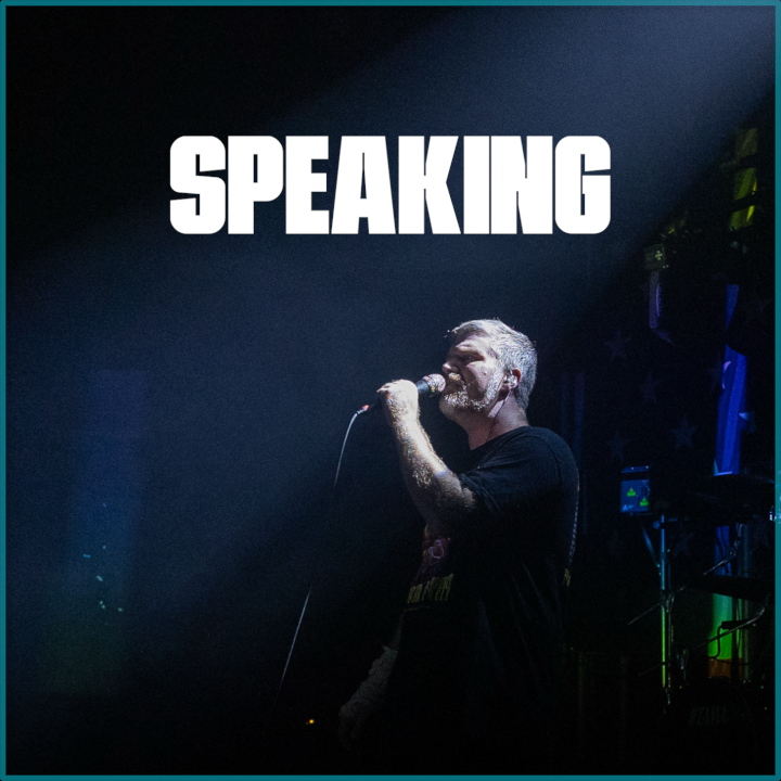 Man speaking into a microphone on stage with the word 'SPEAKING' in large white letters at the top.