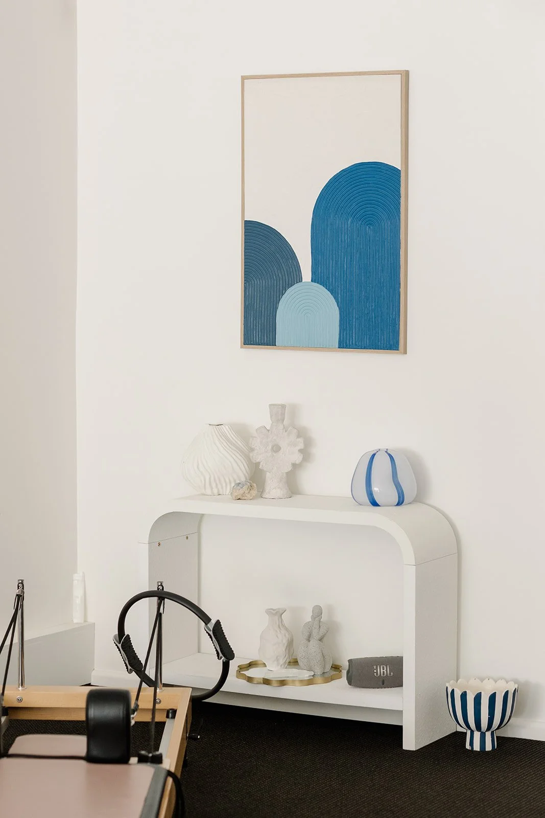 Decorative white console table with vases, sculptures, against a white wall with a blue abstract art piece hanging above. A reformer can be seen in the foreground with a pilates ring.