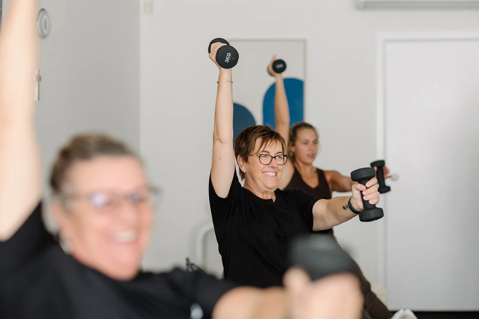 Women exercising with dumbbells in a pilates class, smiling and raising dumbbells overhead.