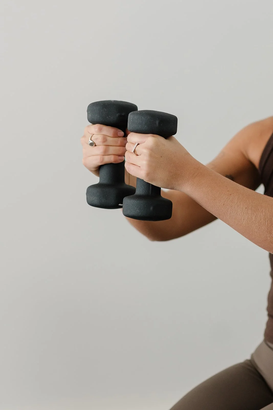 Person holding two black dumbbells in each hand, wearing rings, with a light gray background.
