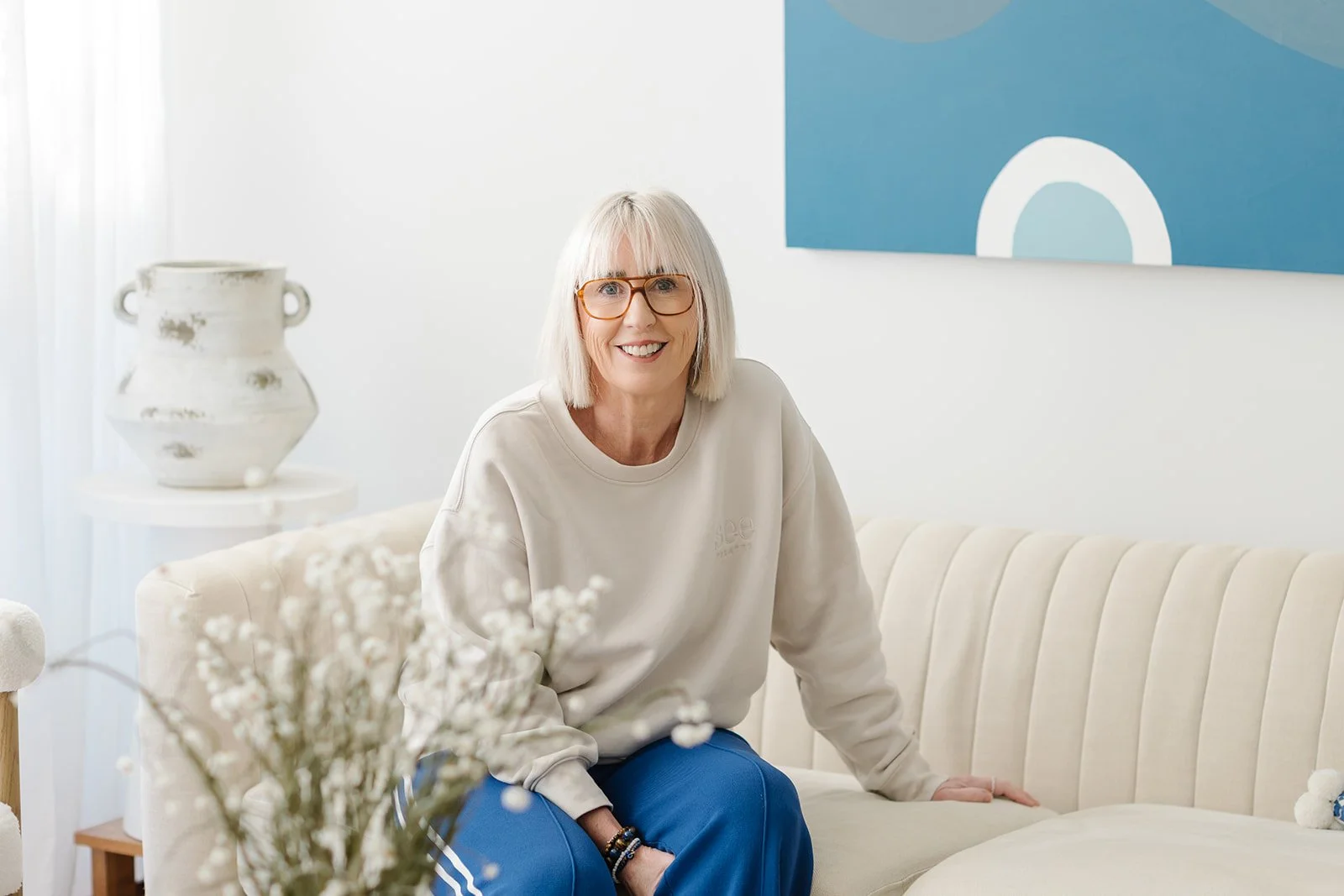 Simone from See Pilates is sitting on a cream sofa in the foyer of the studio, decorated with a white vase and framed blue artwork.