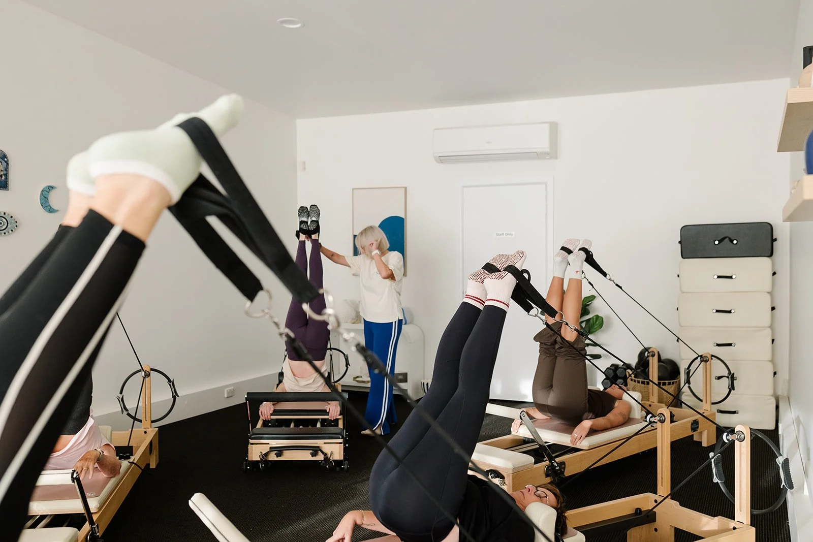 People doing a reformer pilates class with feet in straps. Simone is assisting and checking form.