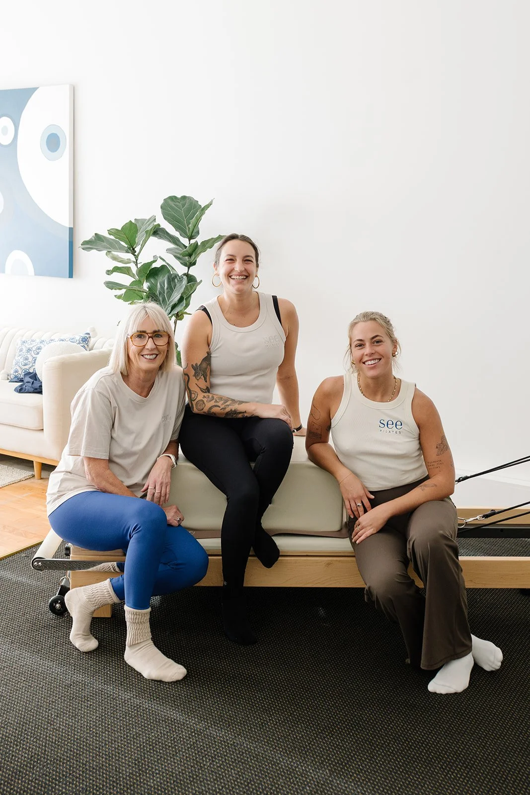 The See Pilates team smiling and sitting on a reformer machine at a See Pilates studio, with a potted plant, abstract wall art, and white walls in the background.