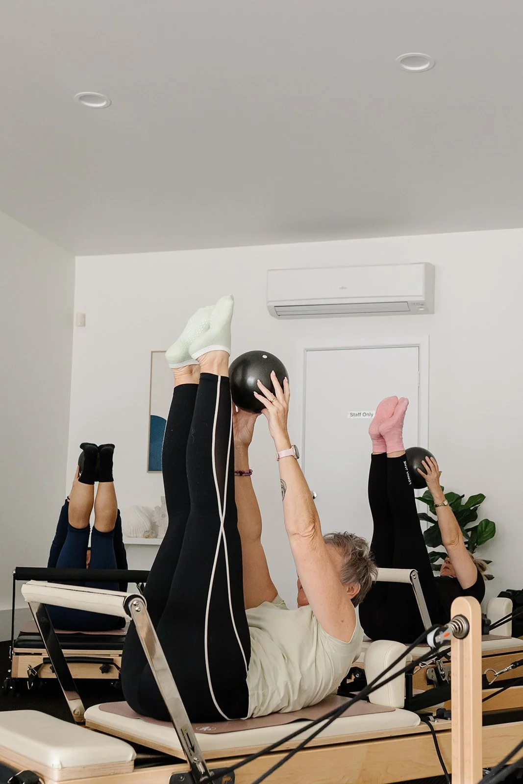 Three women participating in a pilates class in the studio, lying on their backs with legs raised, some wearing socks and leggings.