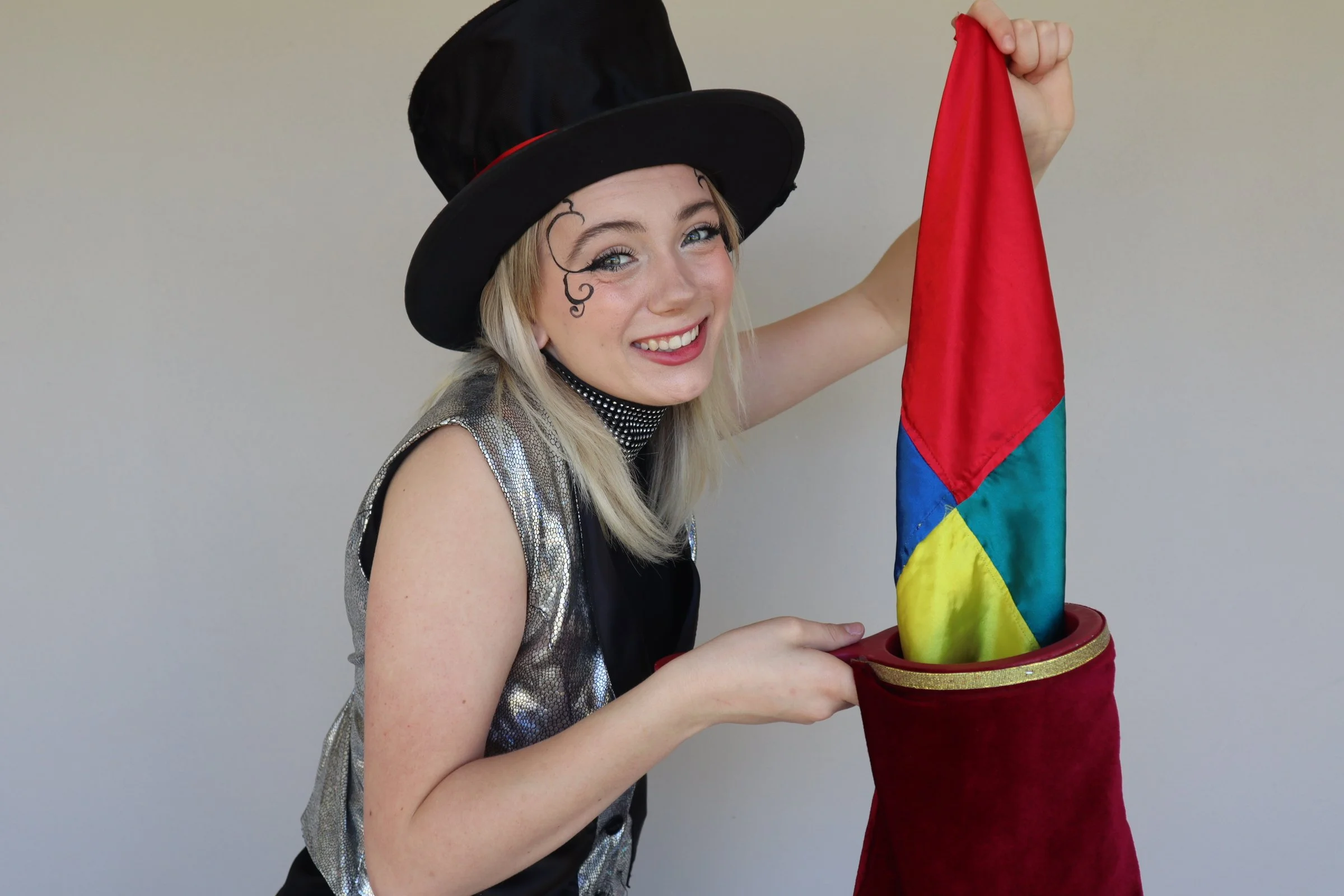 Magic Macy with a black top hat and silver vest, holding a red bag with a rainbow-colored cloth emerging from it, smiling at the camera. Imagination Makes Magic, interactive preschool show.