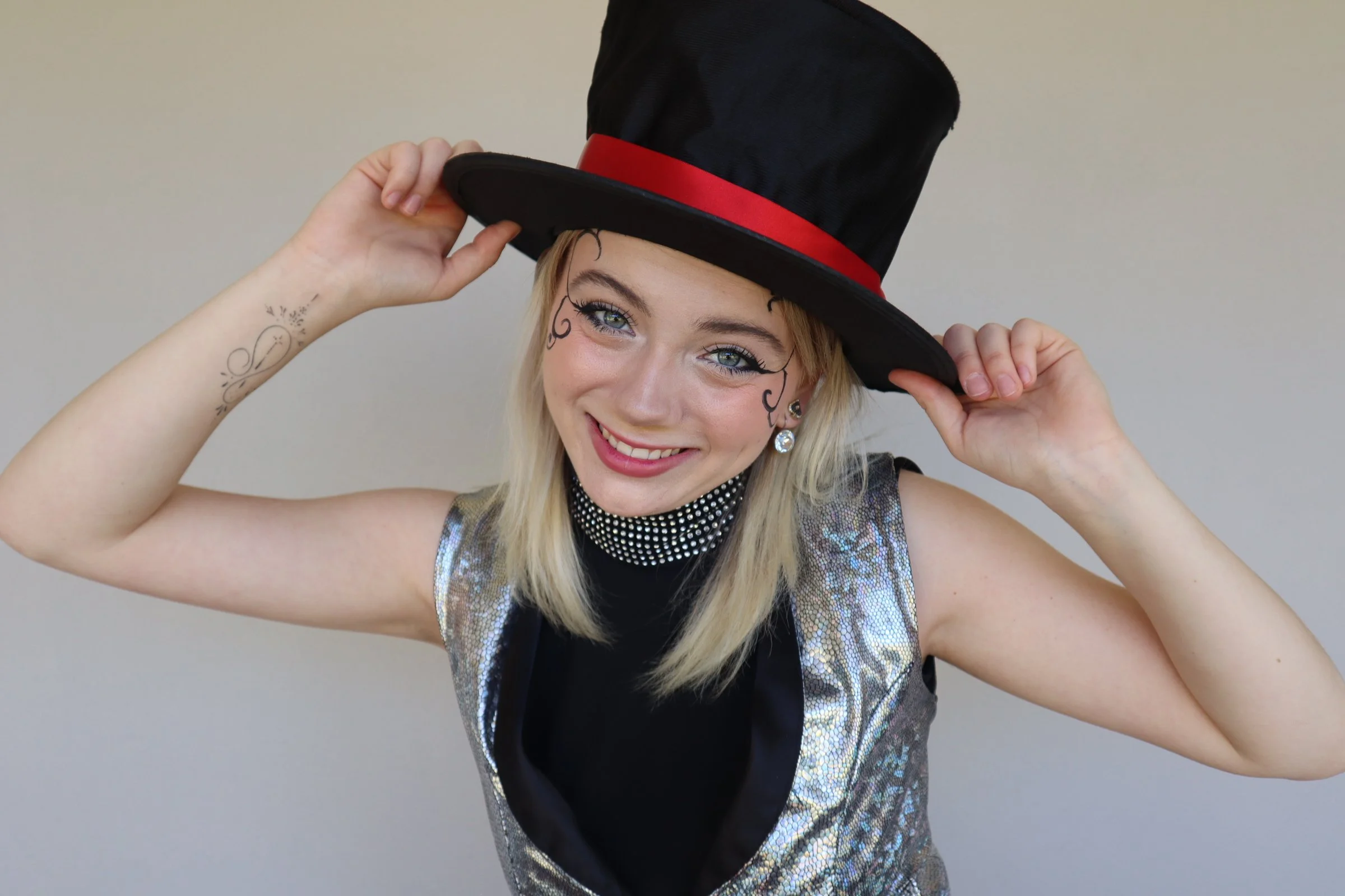 A young woman with blonde hair and tattoos on her arm, smiling and wearing a black top hat with a red band. She is adjusting her hat with both hands, and her face has artistic black face paint design elements around her eyes. Magic Macy.