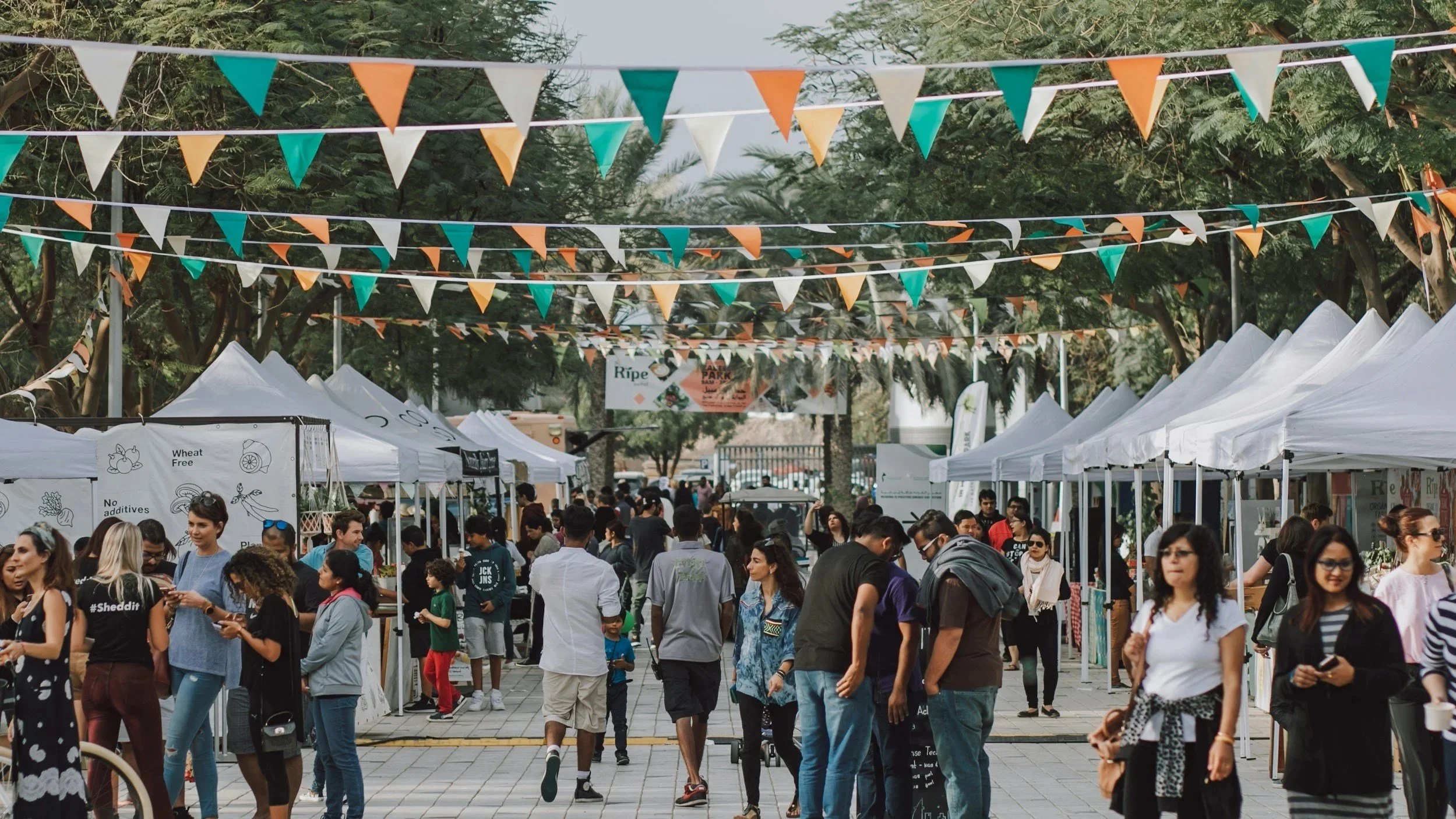 Outdoor community market with people walking between vendor booths and gathering in a social public space.
