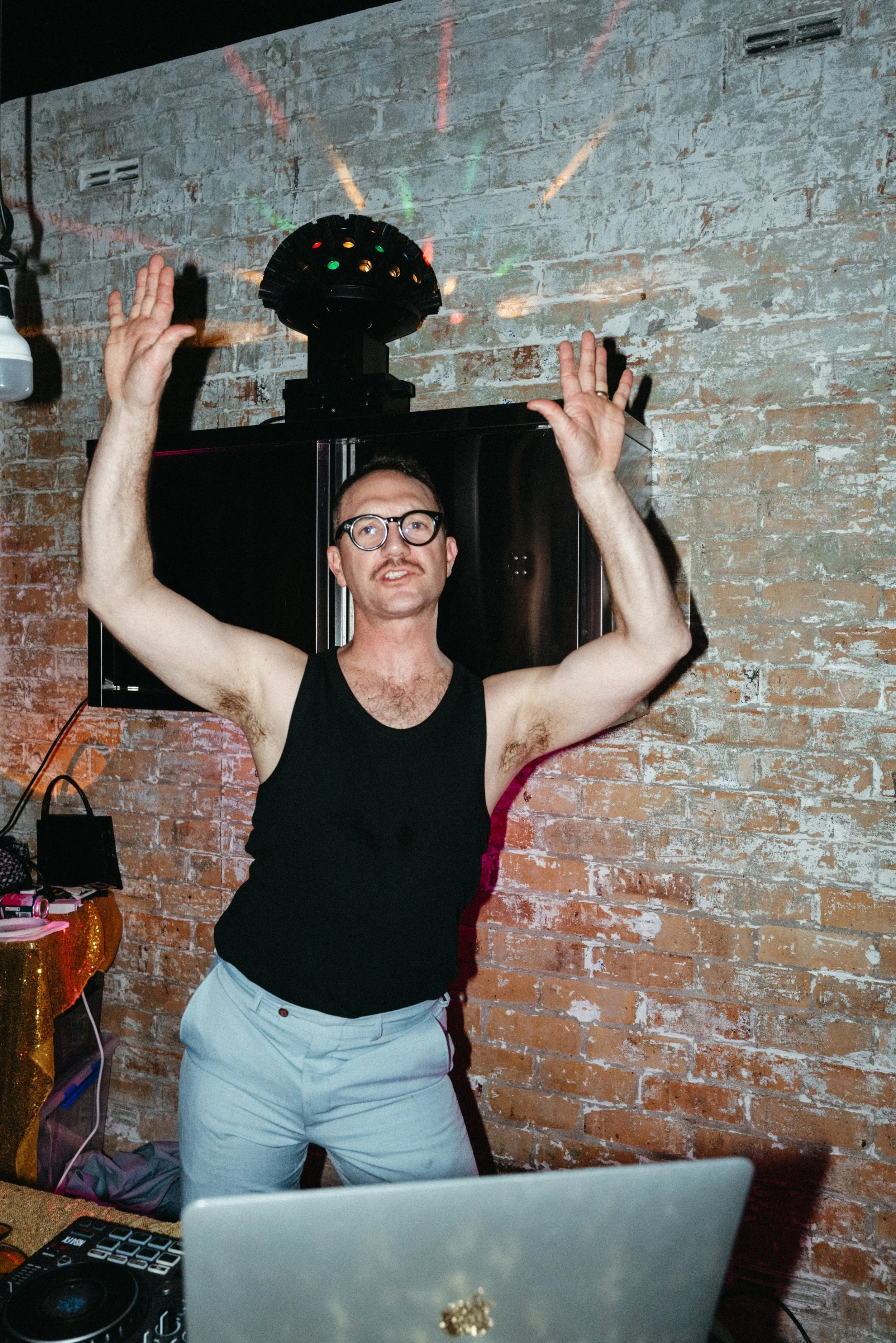 A man wearing glasses, a black tank top, and light-colored pants dancing with his arms raised in front of a brick wall. There is a disco ball with colorful lights on the ceiling above him.