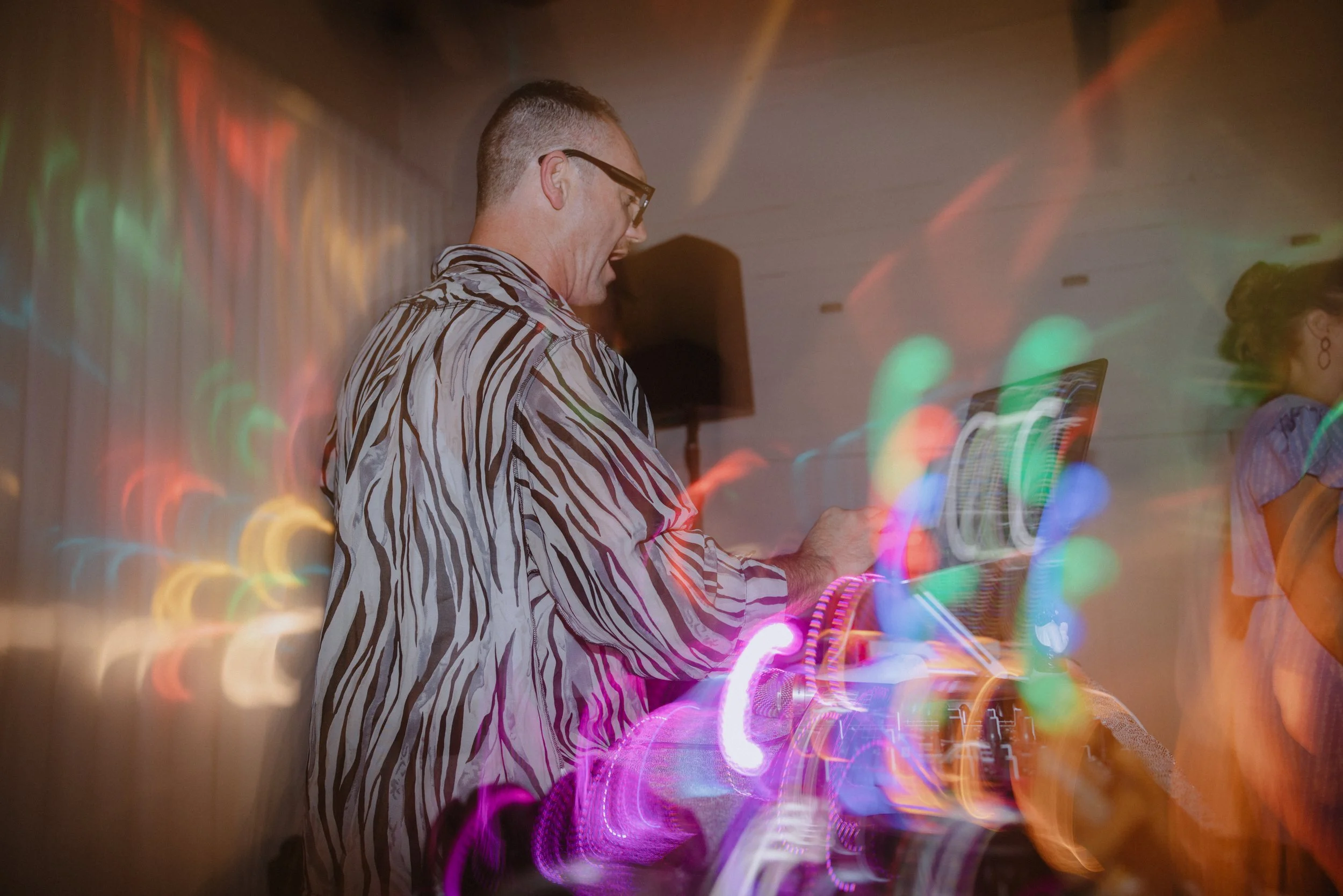 A man wearing glasses and a zebra-striped shirt is DJing at an event with colorful light effects.