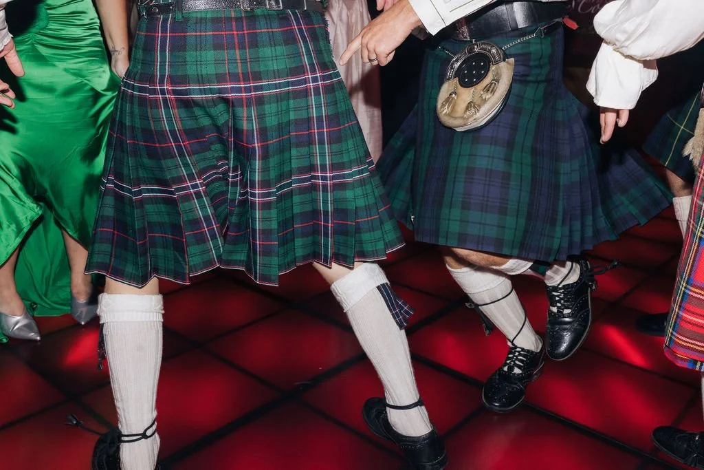 People wearing traditional Scottish kilts and knee-high socks dancing on a red-lit floor.