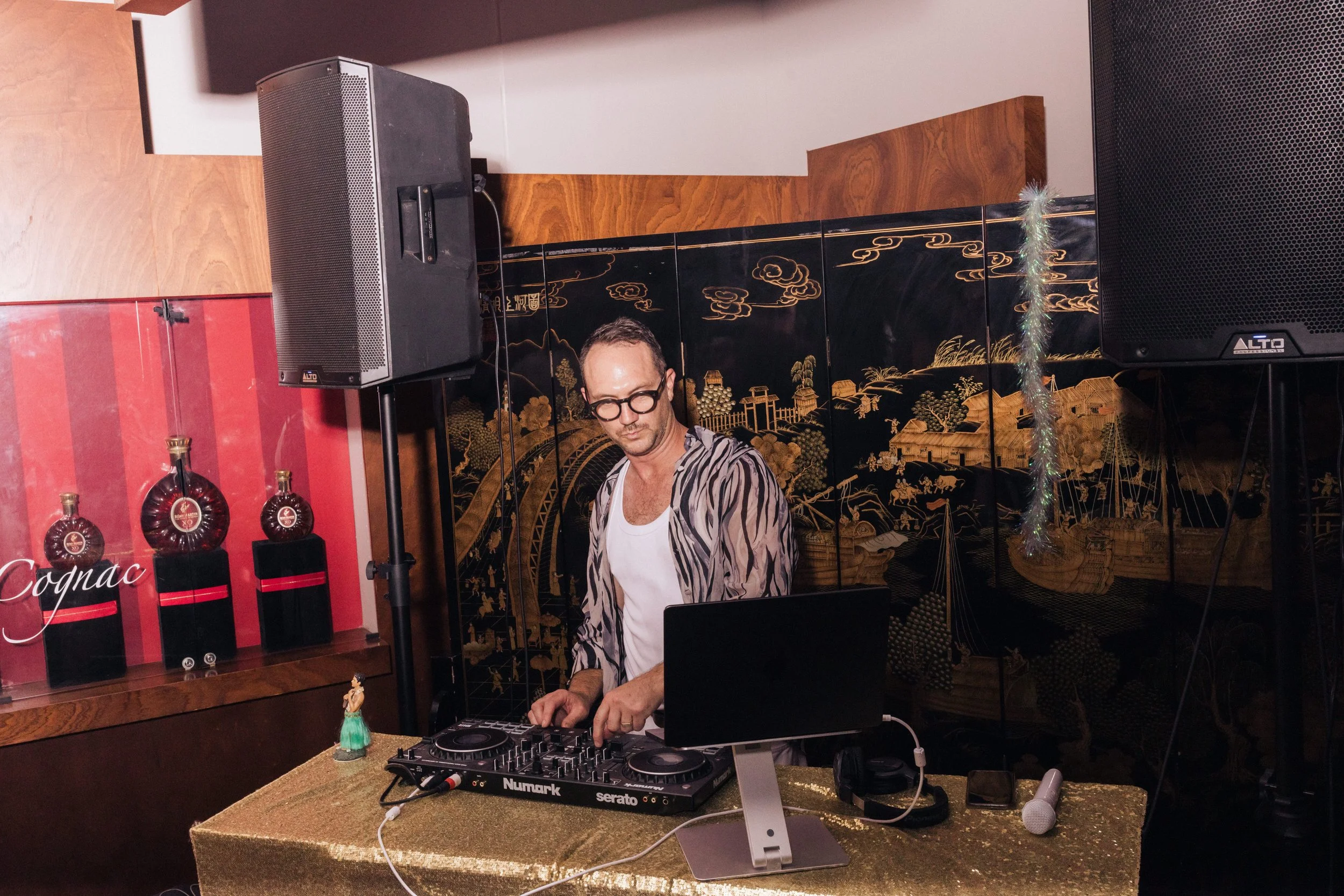 A man DJing at an event, with DJ equipment, speakers, and decorative Asian-style background.