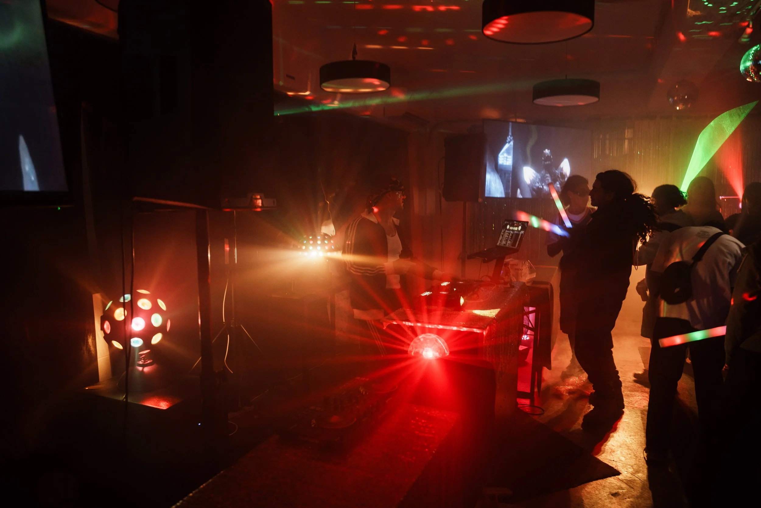 People dancing at a party with colorful lights, DJ equipment, and a large screen displaying a black-and-white video.