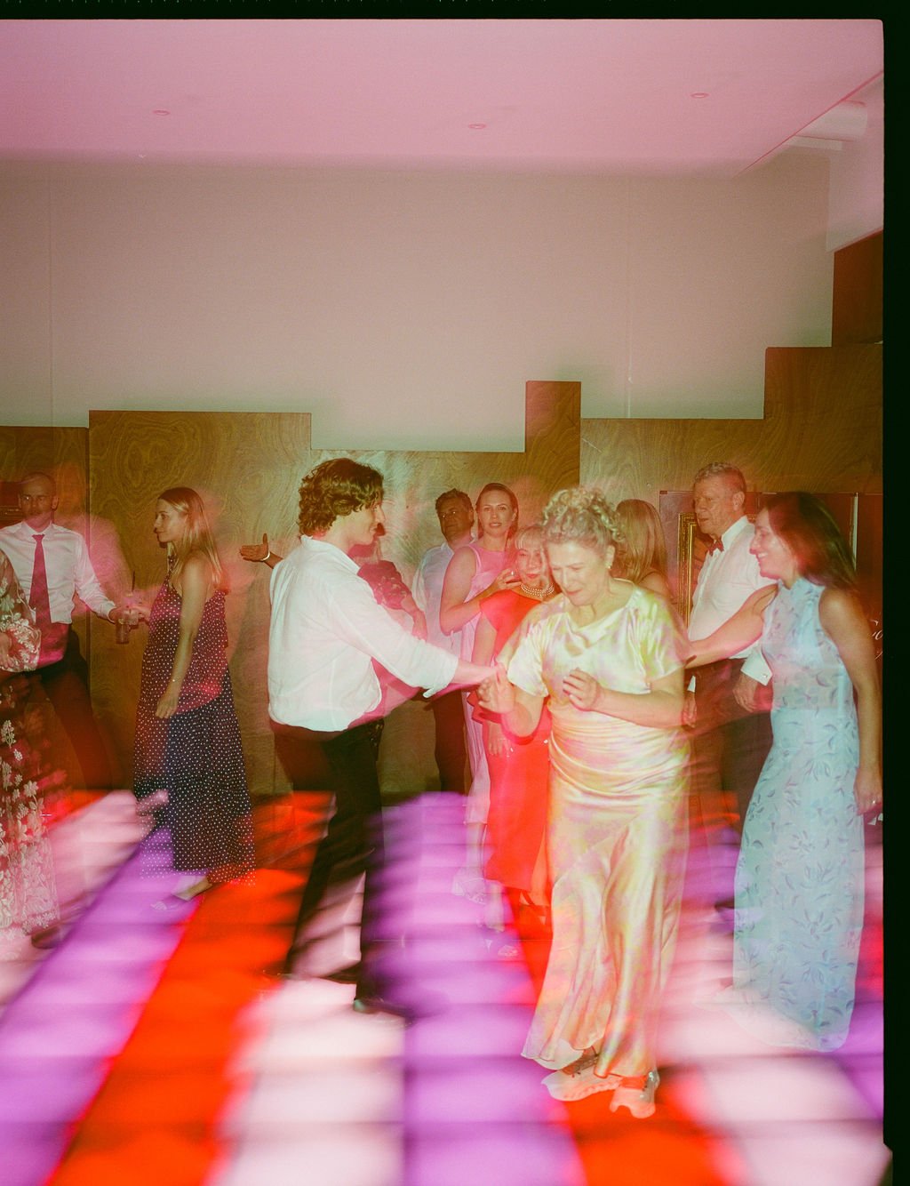 People dancing and socializing at a party or celebration in a room with wooden walls and colorful lighting.