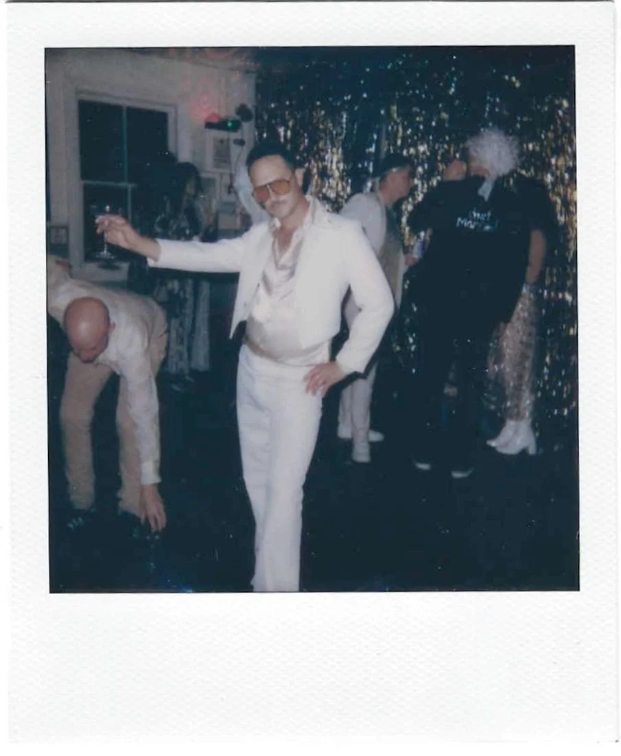 Man dressed in a white tuxedo with glasses and a headband, holding a drink, posing in front of a shiny, gold party backdrop with other people in fancy outfits behind him.