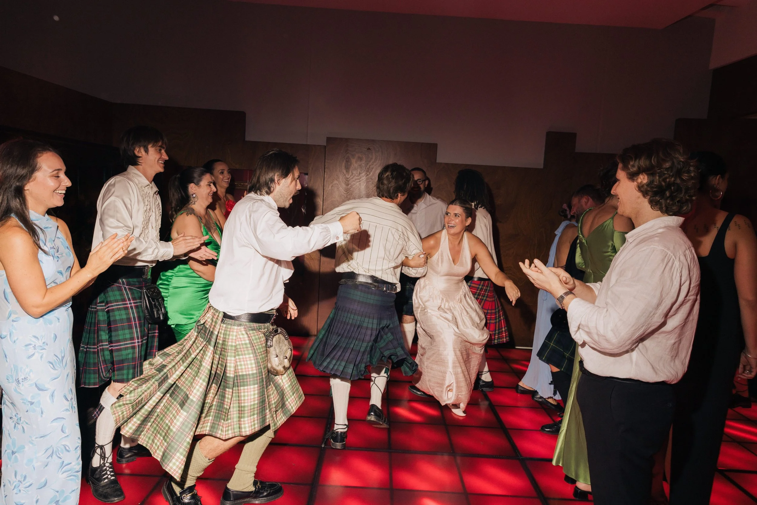 People dancing and celebrating at a party, some wearing kilts, on a red-lit dance floor.