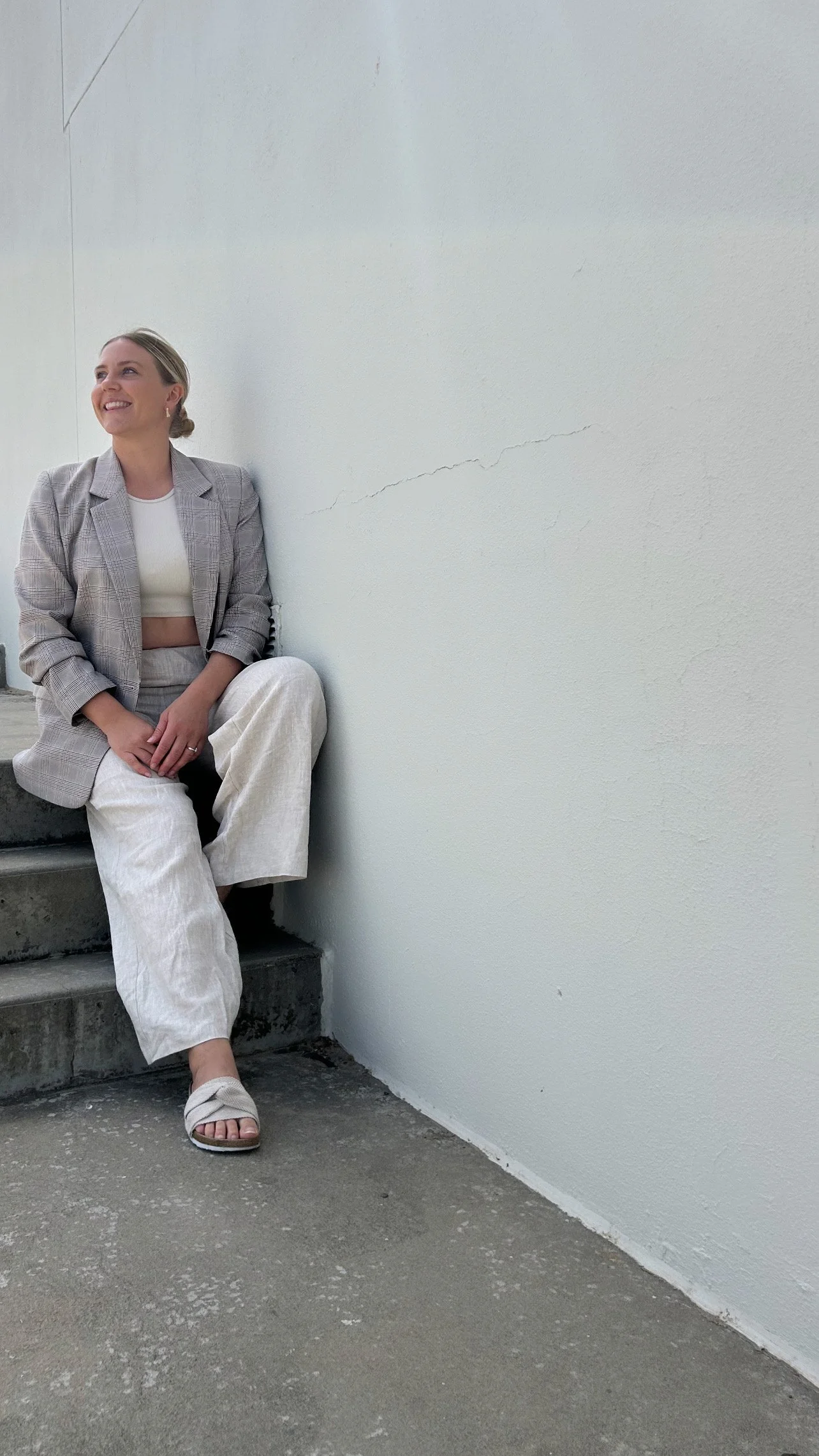 Person sitting on steps against a plain wall, wearing a plaid blazer, white crop top, light-colored pants, and sandals, looking upwards and smiling.
