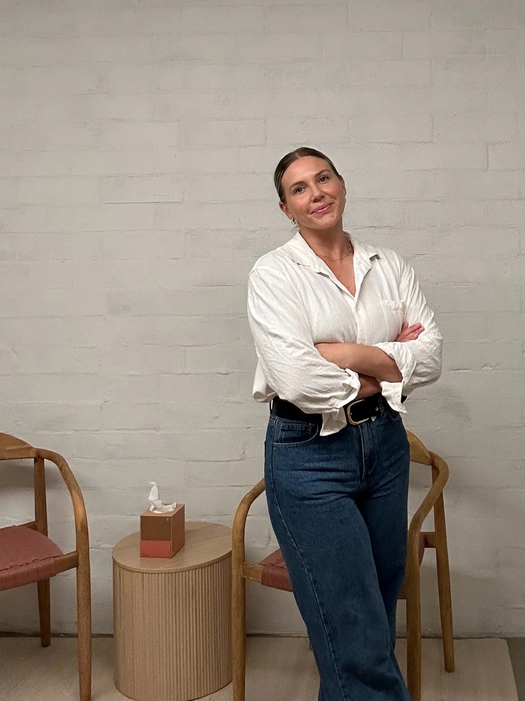 Person wearing a white shirt and blue jeans with arms crossed, standing in a room with two wooden chairs and a small table with a tissue box.