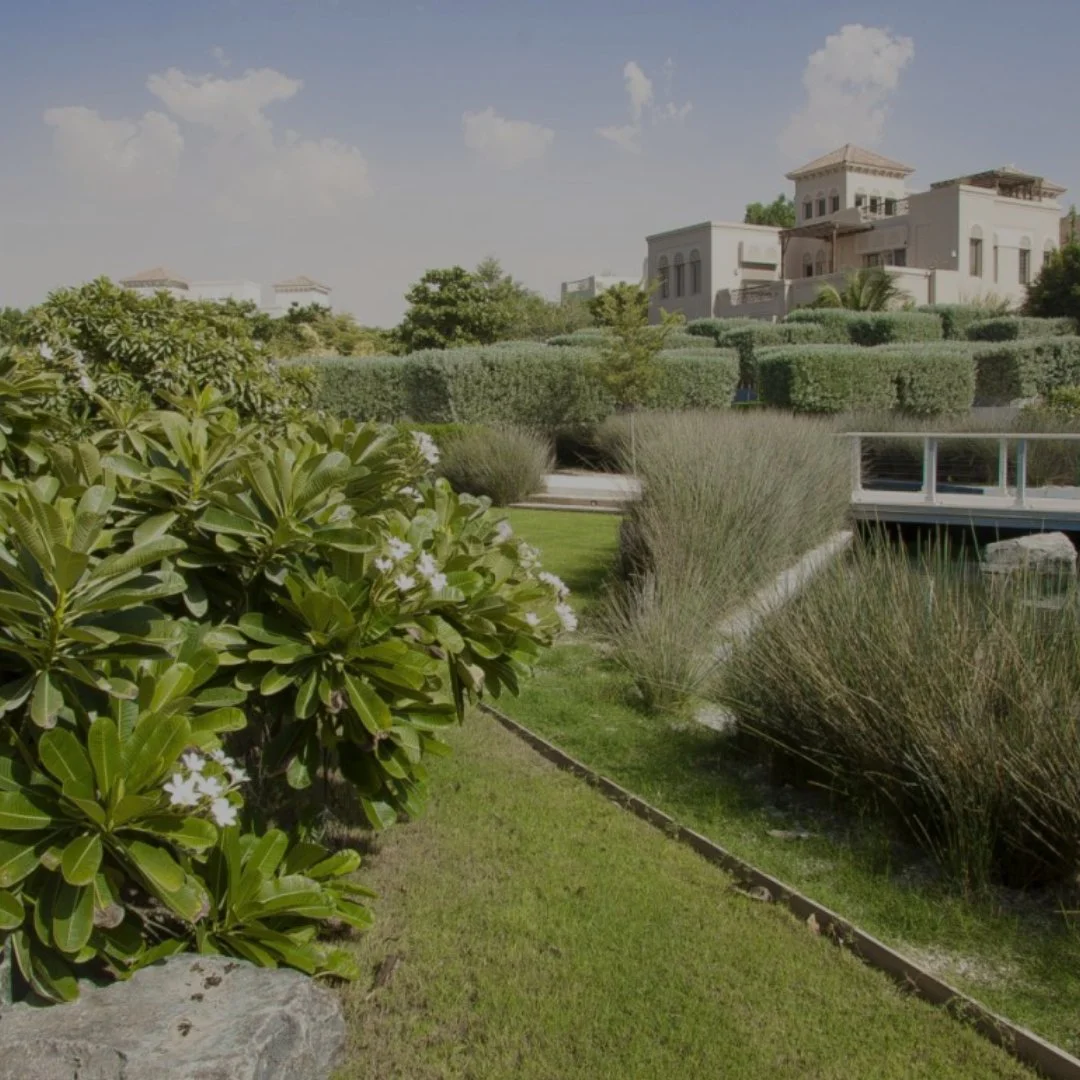 A landscaped garden with trimmed bushes, tall grasses, and a pathway in front of residential buildings with multiple stories and terraces. Clear sky with some clouds.