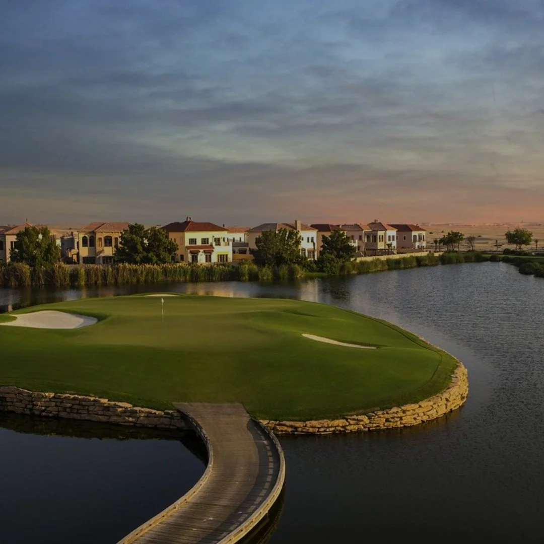 A golf course green with sand traps and a flag, overlooking a water body with a curved stone edge, with residential houses in the background and a cloudy sky at sunset.