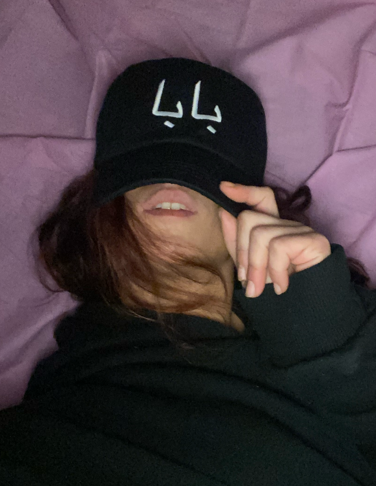 Person lying on a bed, wearing a black cap with white Arabic script, covering part of their face with their hand, and wearing a black hoodie, with purple sheets in the background.