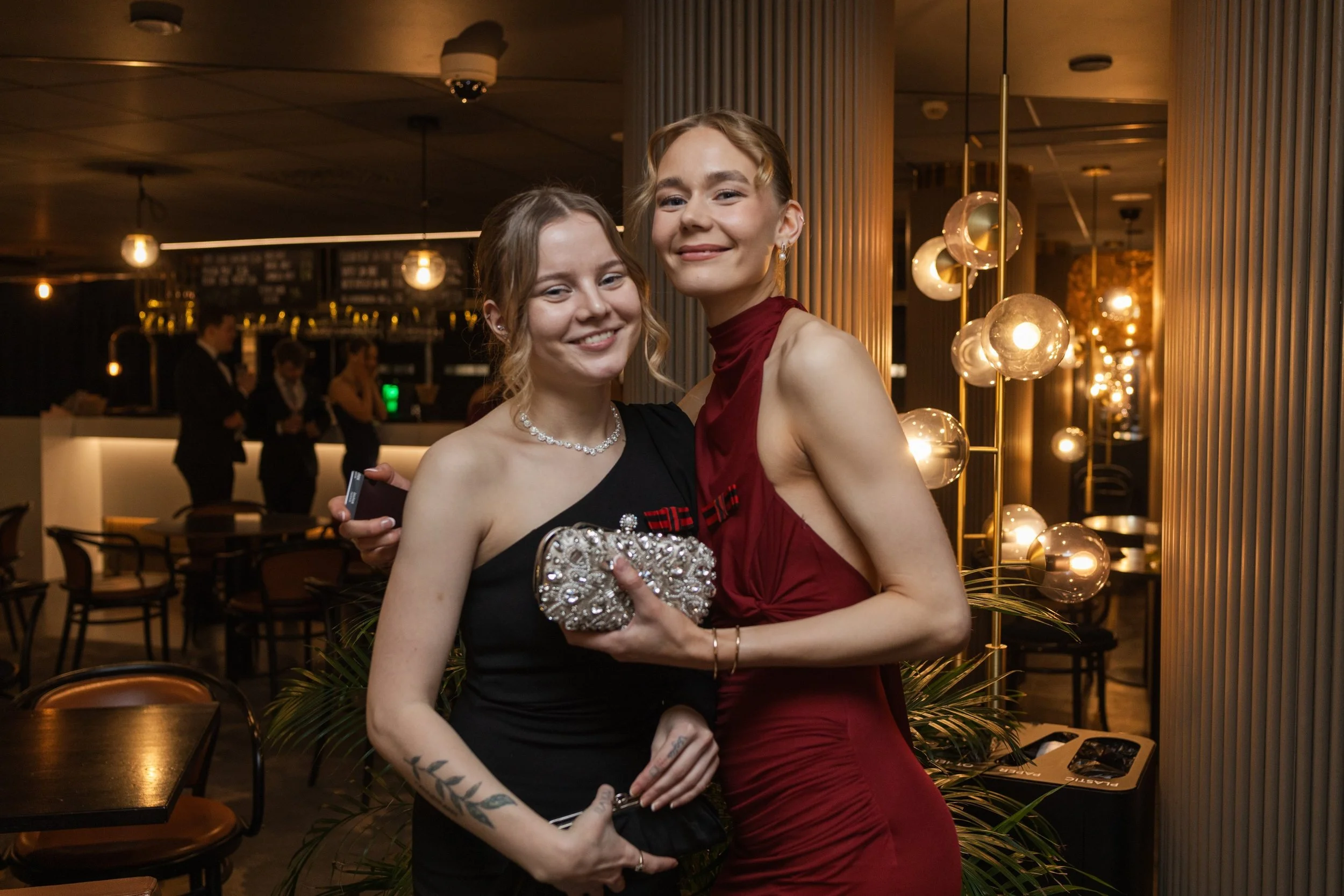 Kaksi nuorta naista hymyilemässä ja poseeraamassa juhlassa, yksi pitää riipuslaukkua ja toinen piilottaa jotain kädessään, taustalla ravintola tai juhlatila.