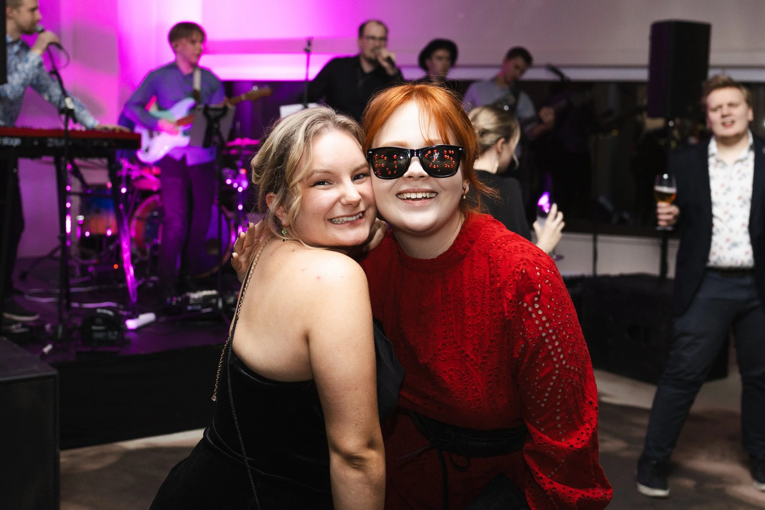Kaksi naista hymyilevät ja poseeraavat tanssiaisissa, taustalla live-musiikkibändi ja muita ihmisiä juovana ja katsomassa.