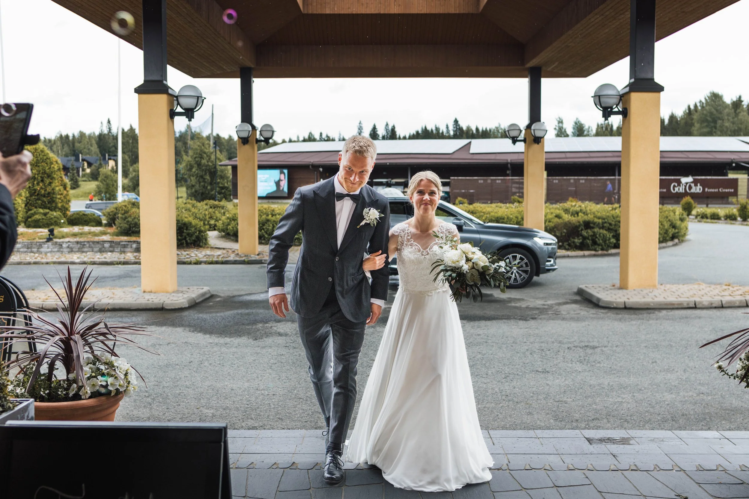 Hääparin kävely ulkona, nainen valkoisessa hääpuvussa ja mies pukussa, taustalla auto ja rakennus, kyltit näkyvissä.