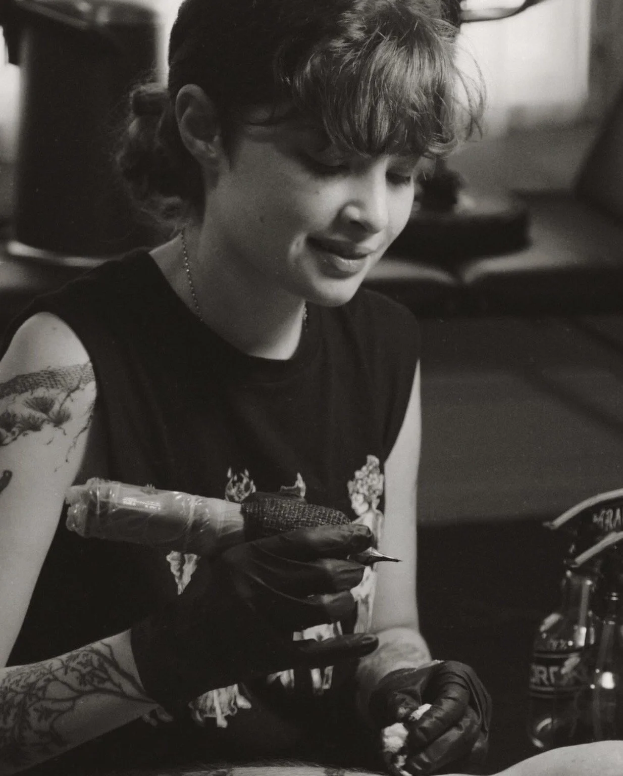 A woman with short brown hair, tattoos on her arms and legs, sitting cross-legged on the tile floor, smiling at a large brown dog sitting next to her by a glass sliding door in a dimly lit room.