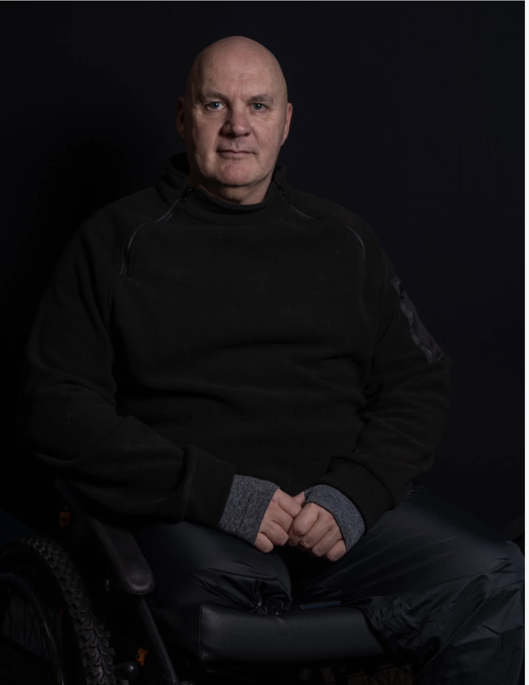 Portrait of a middle-aged man with a bald head, sitting in a wheelchair, wearing a black jacket and gray cuffs, against a dark background.
