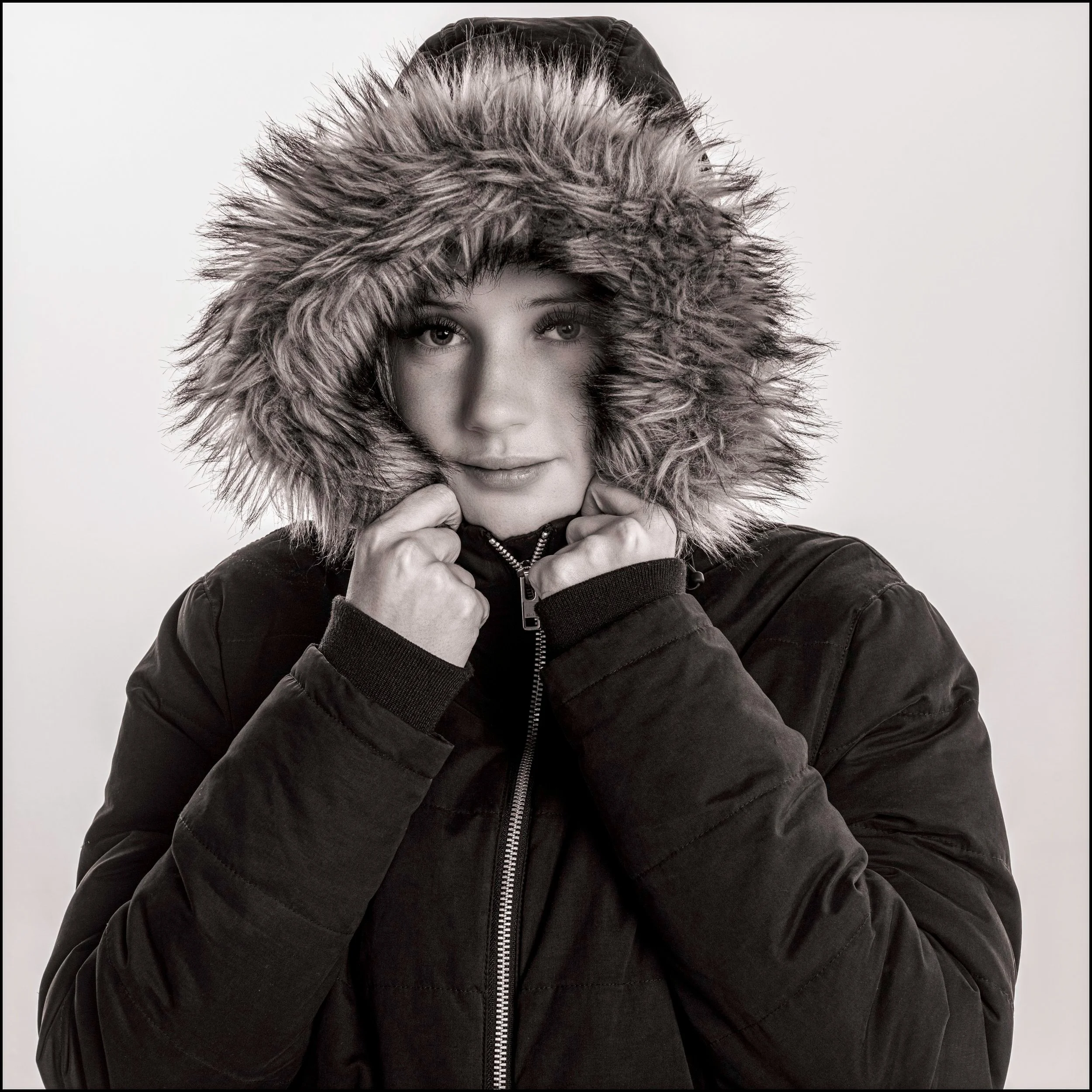 Person wearing a fur-lined hooded jacket, looking directly at the camera, black and white photo.