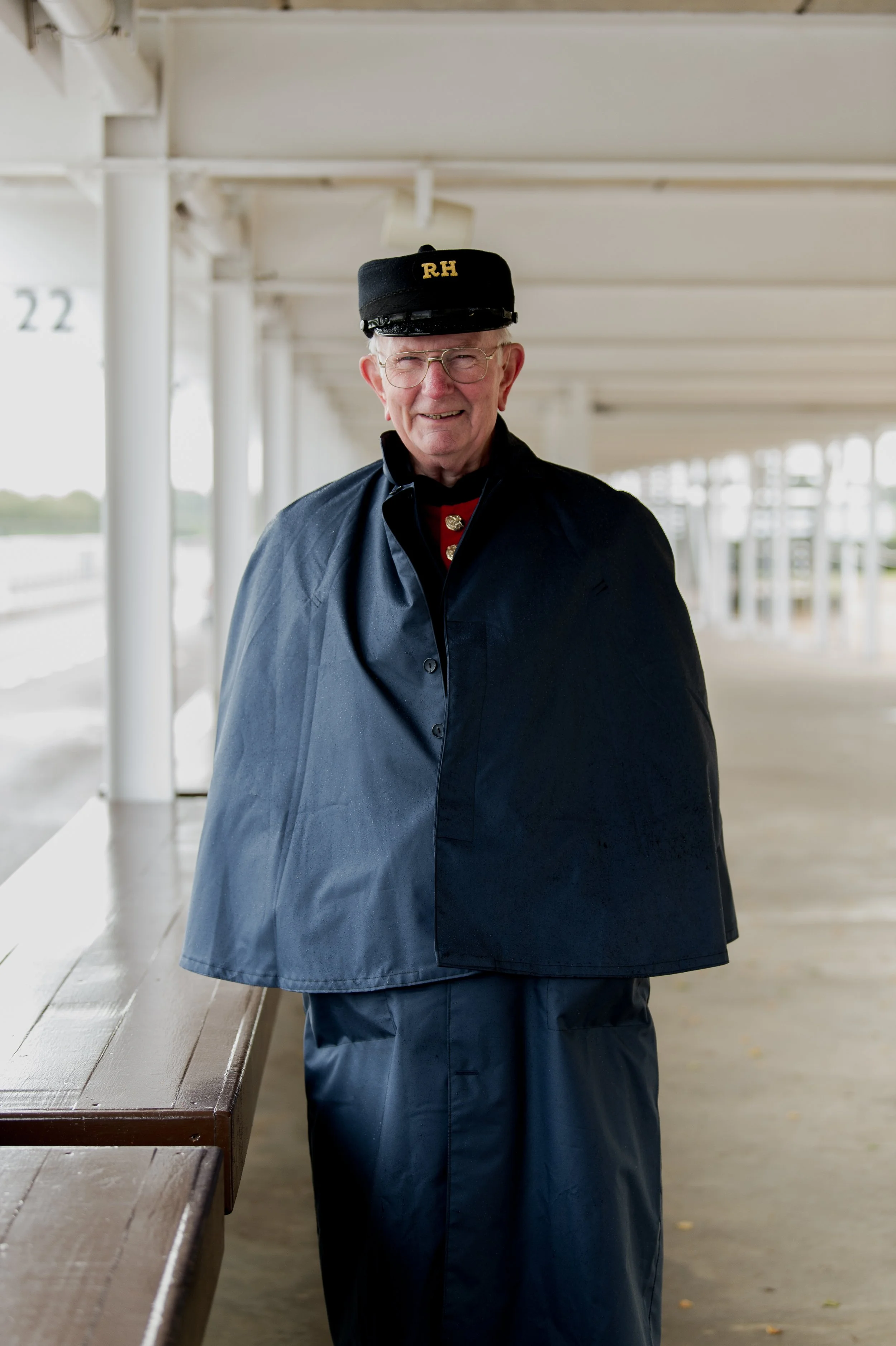 Elderly man in navy blue uniform with 'RH' on cap, standing in covered walkway.