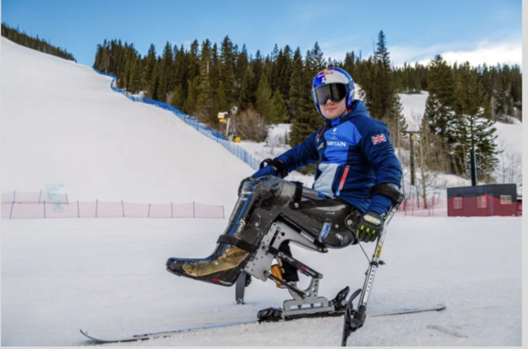 A person in a blue jacket and helmet sitting on a ski chairlift or adaptive skiing equipment on snowy terrain at a ski resort, with pine trees and ski slopes in the background.