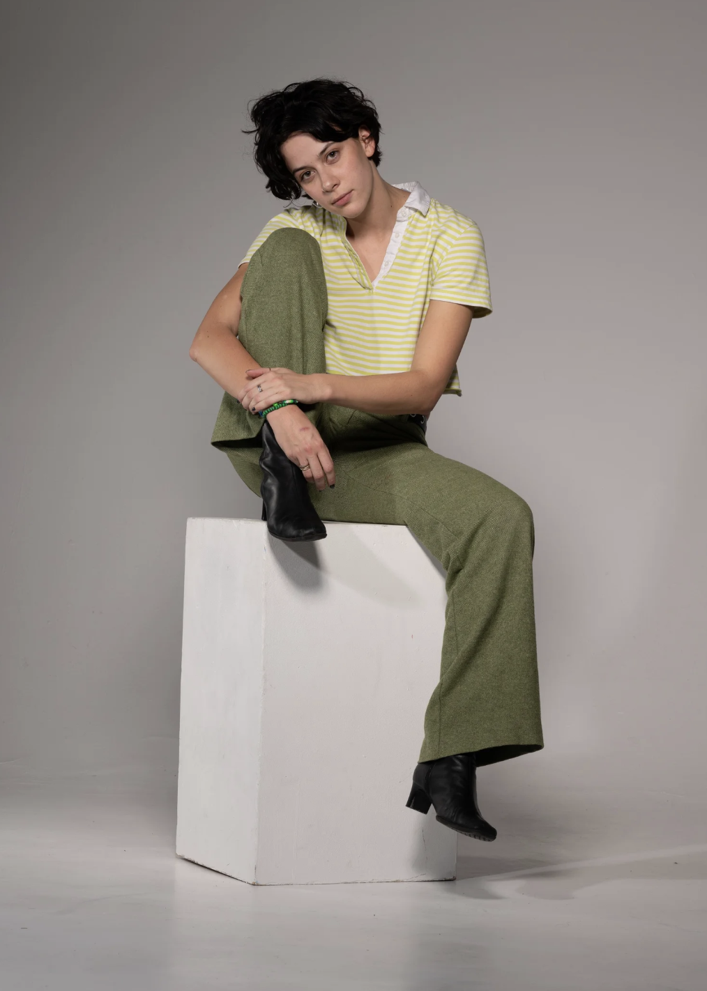 A young woman with short black hair, wearing a yellow striped short-sleeve shirt and green wide-leg pants, sits on a white block in a studio setting with a plain gray background. She has one leg up, her right foot resting on her left knee, and is wea