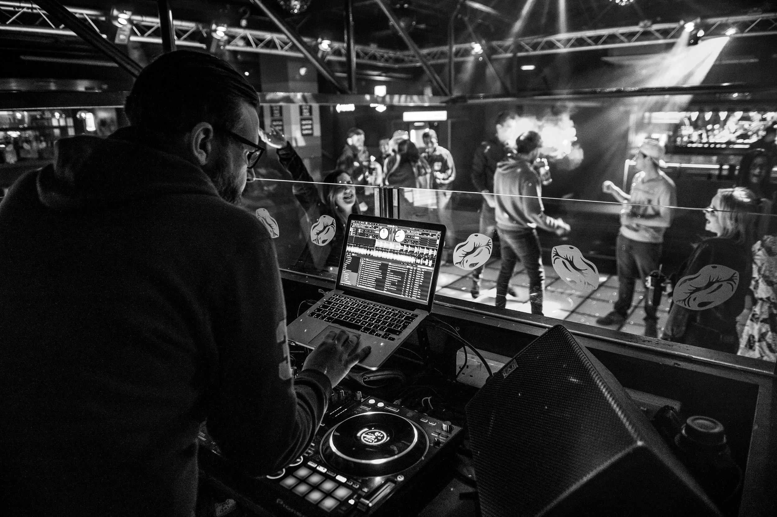 DJ performing in a nightclub with a laptop and turntable setup, visible through a booth window. People are dancing and socializing on the dance floor in the background.