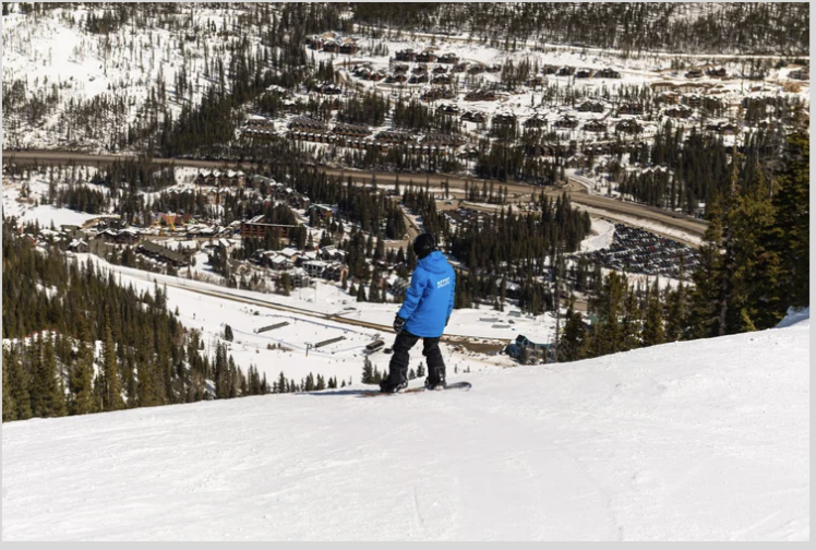 A person in a blue jacket snowboarding down a snowy slope with a town and forested mountains in the background.